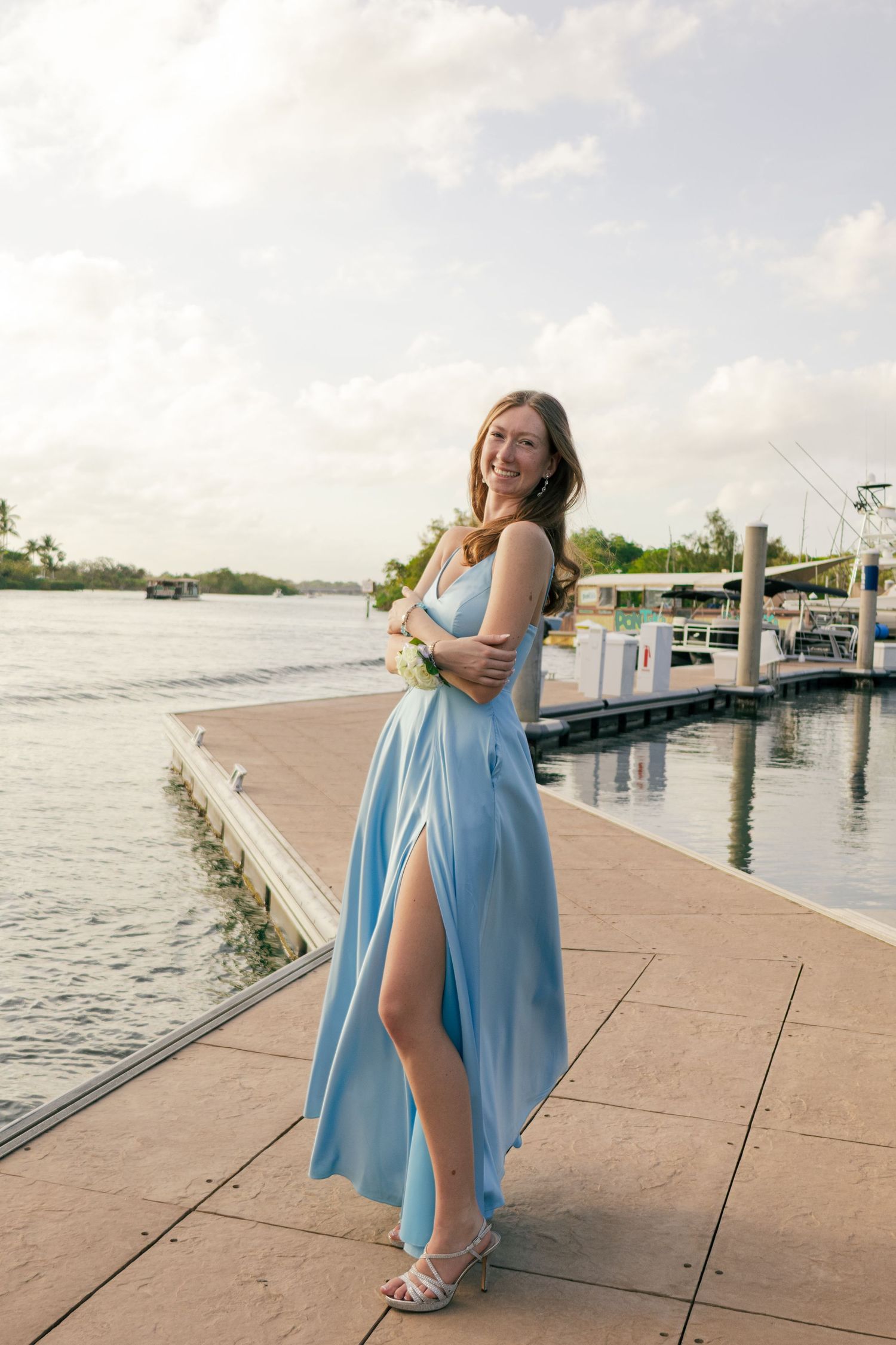 A smiling teenage girl in a light blue satin prom dress holds a corsage on a sunny Florida waterfront dock.