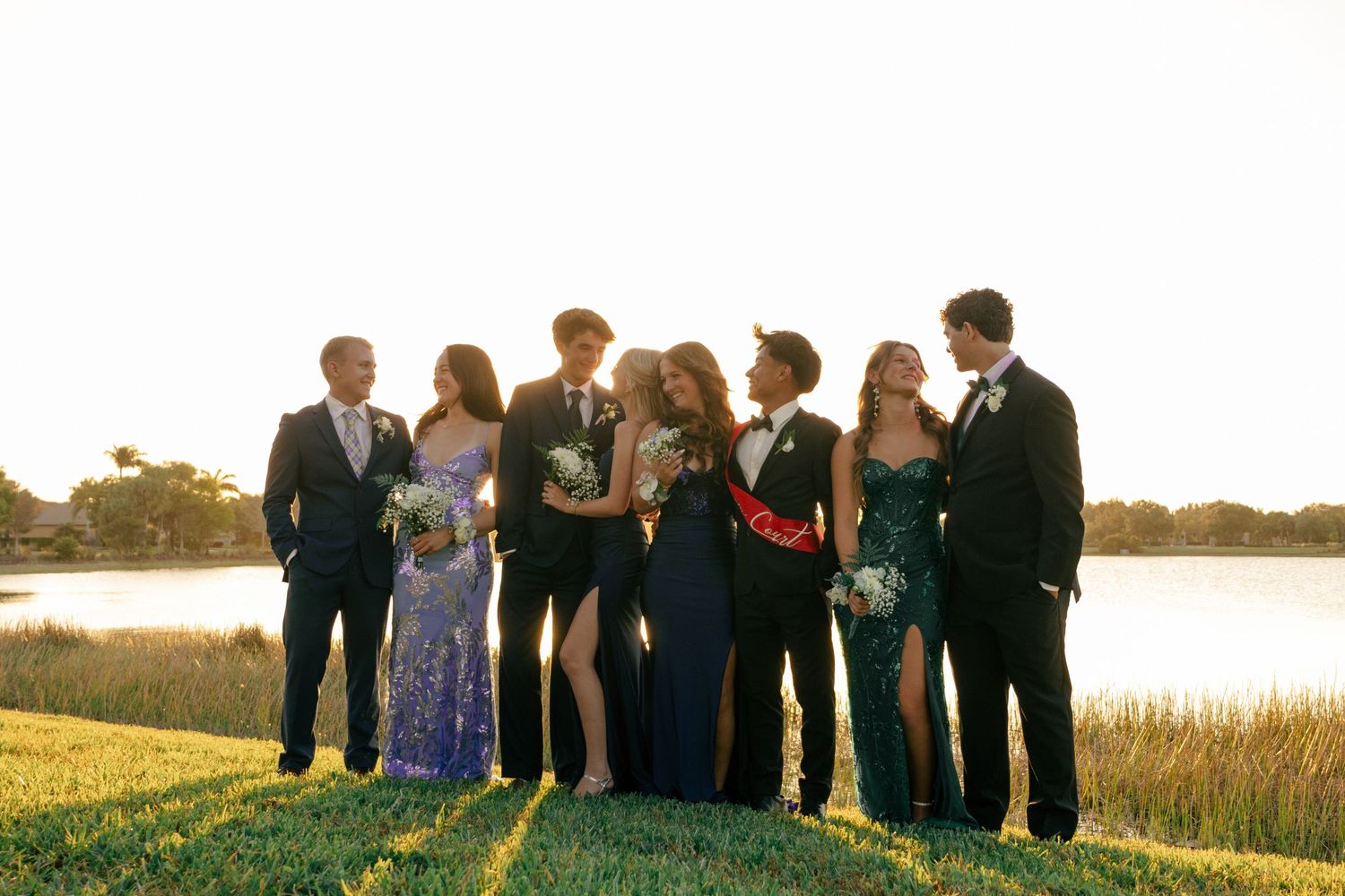 Group of teens in formal prom attire posing outdoors near a lake at golden hour sunset.
