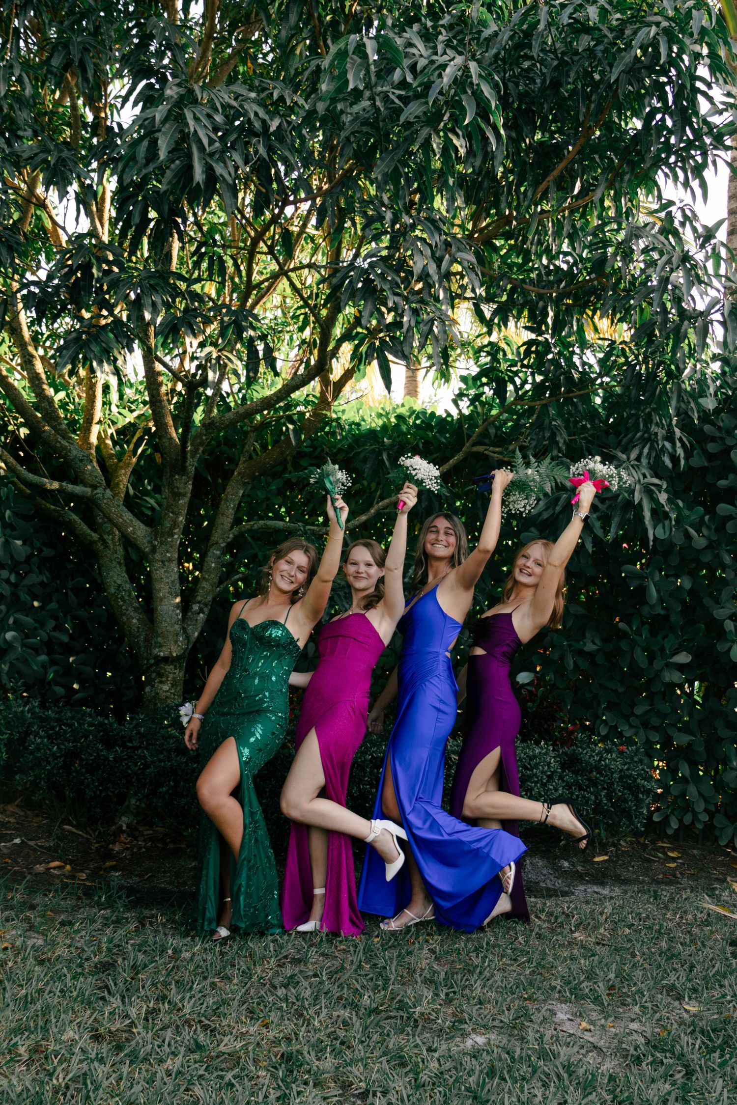 Four young women in vibrant prom dresses jump and celebrate joyfully under a large green tree.
