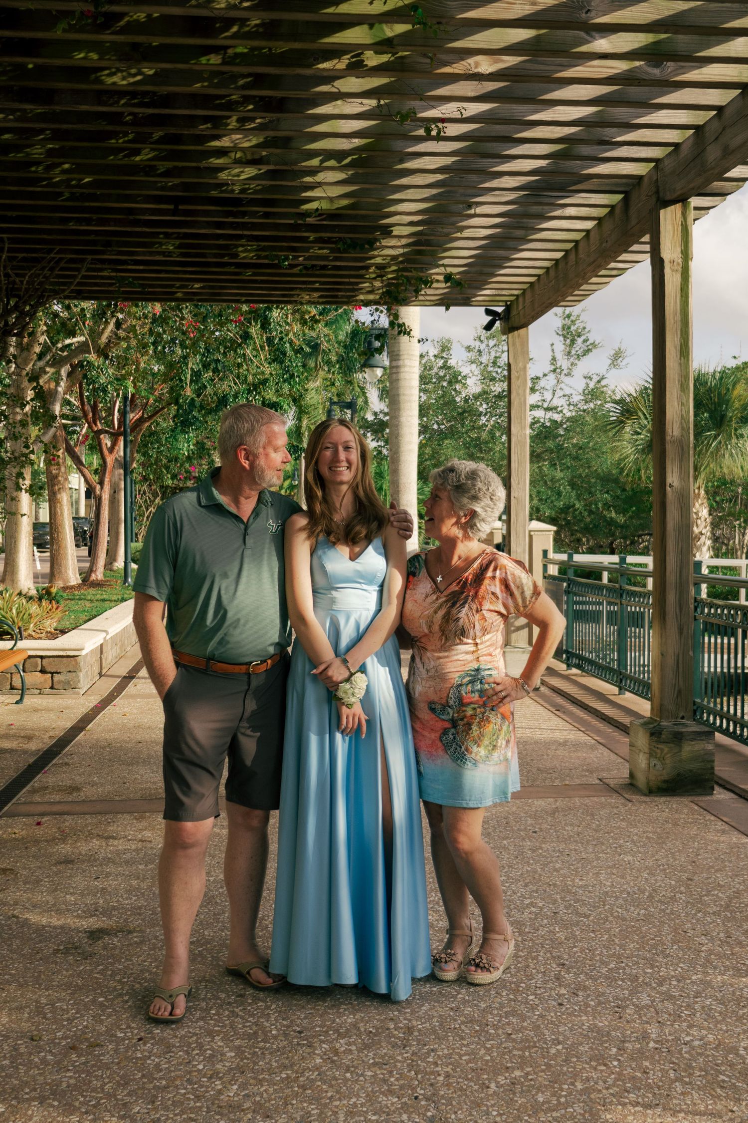 Young woman in a blue formal dress laughing with two adults under a lush outdoor pergola.