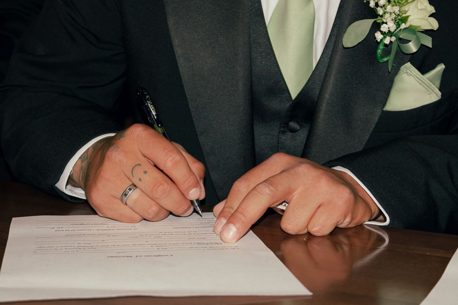 Groom in dark suit signing wedding certificate with boutonniere and ring visible.