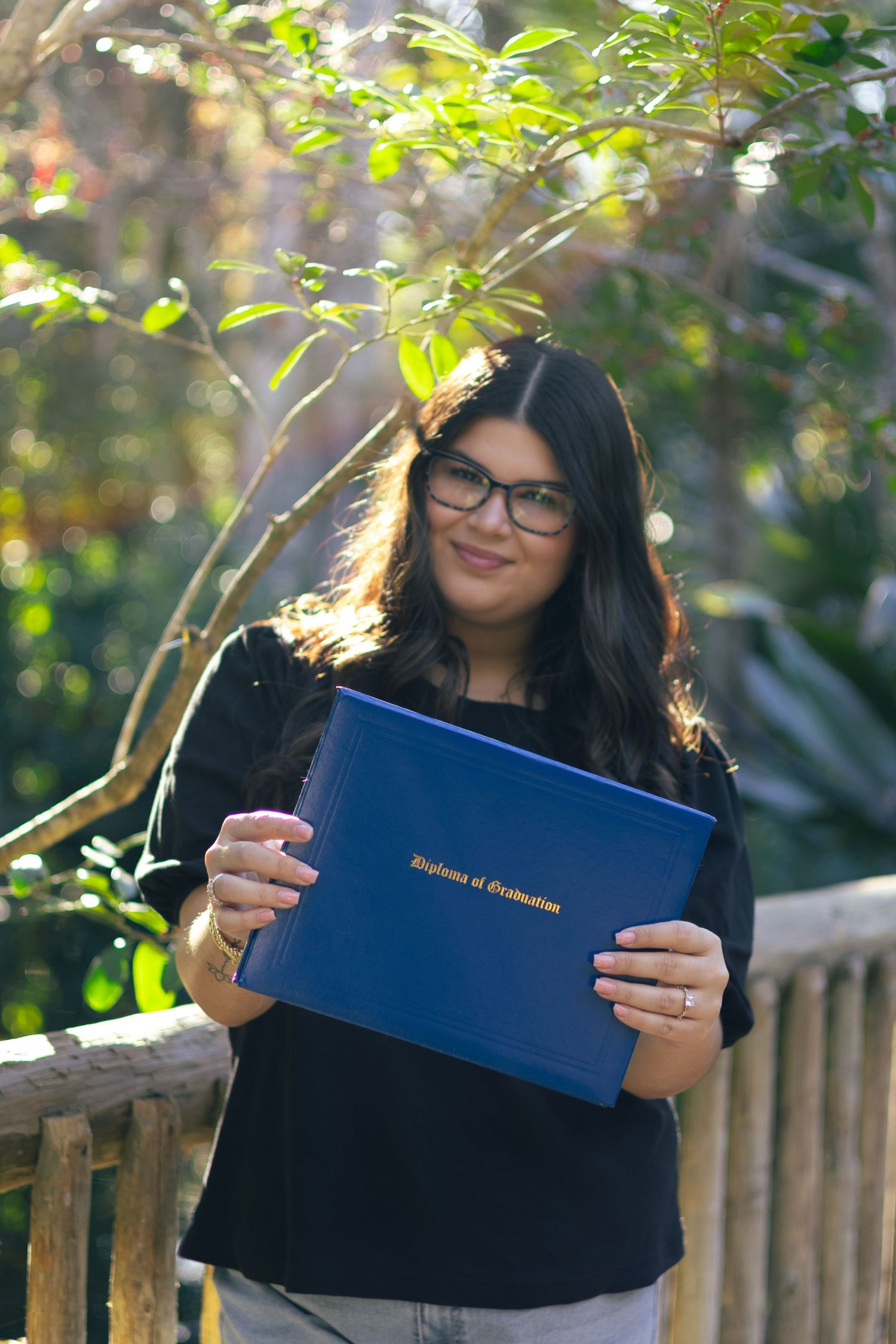 Smiling graduate holding blue Diploma of Graduation cover outdoors in sunlit garden setting.