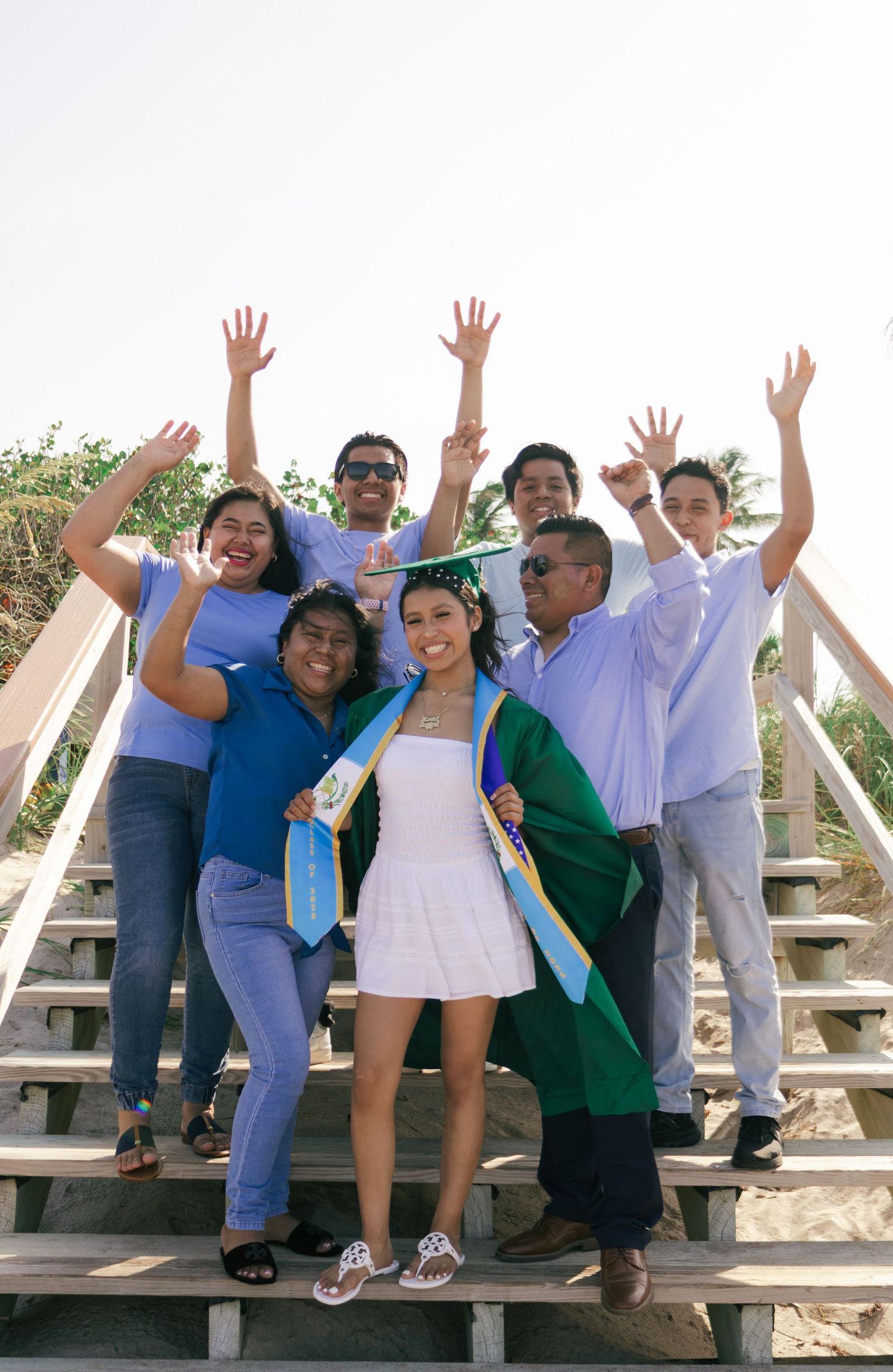 Group of seven people celebrating a graduation on wooden beach stairs, with arms raised in joy, graduate wearing green cap and gown.