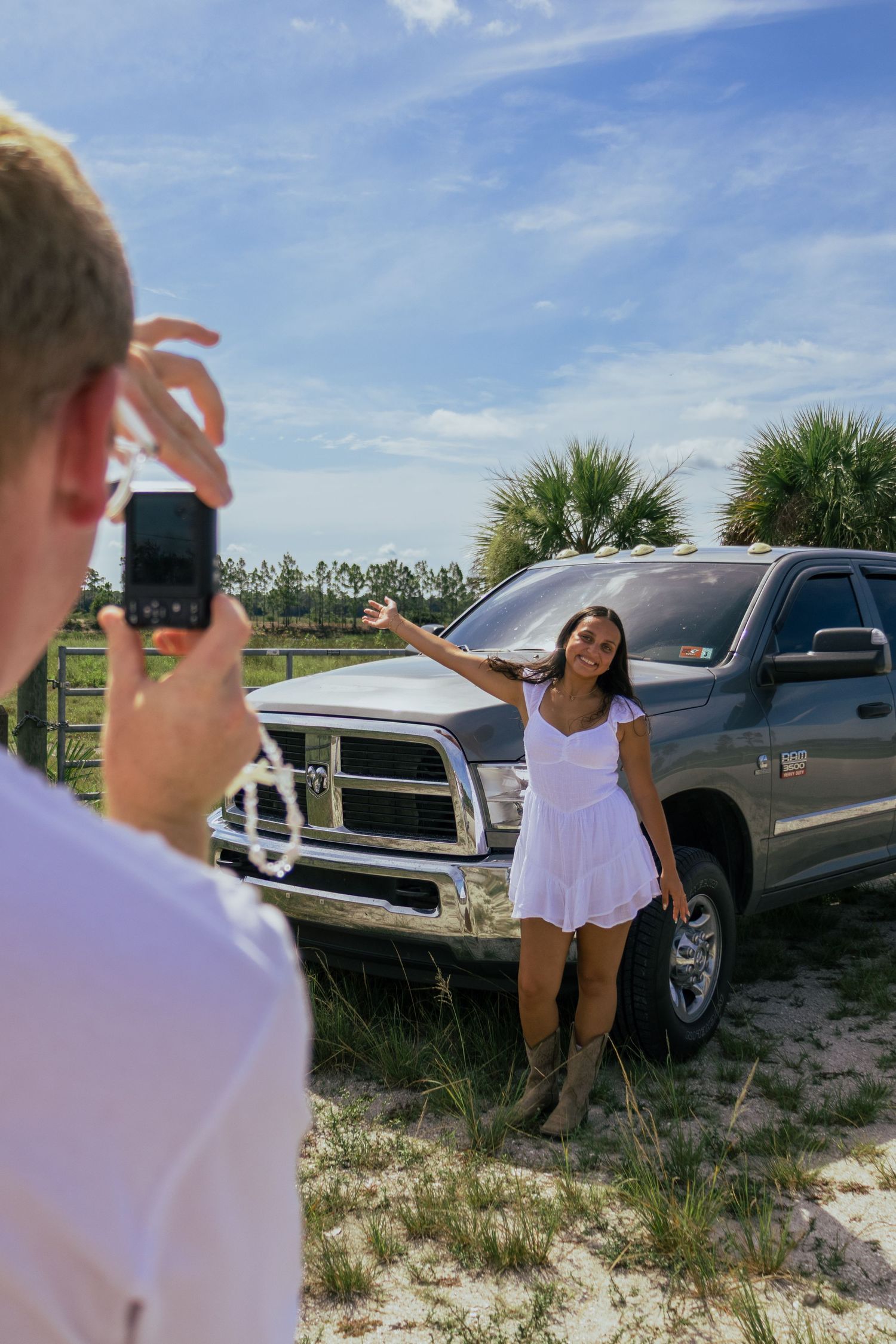 Woman in white dress posing beside a Dodge Ram truck while her partner photographs her outdoors.