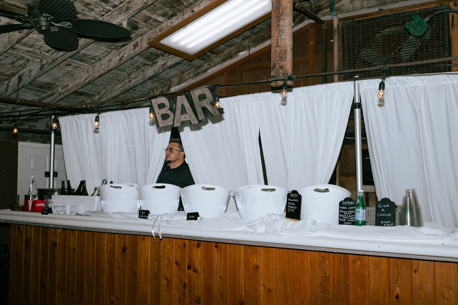 Bartender stands behind rustic wooden bar with white draping and illuminated BAR sign.