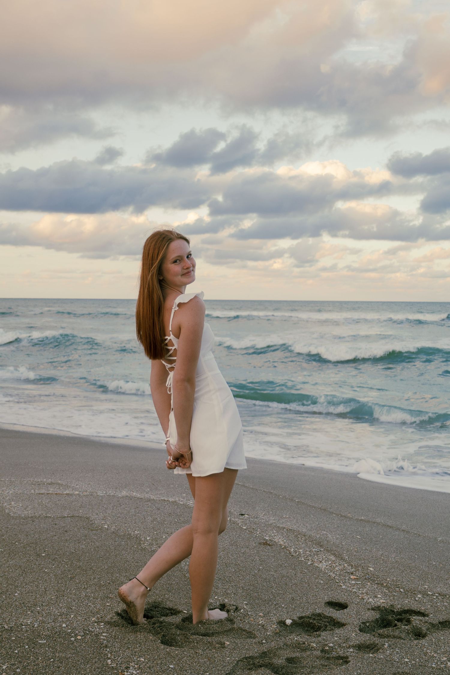 Young woman in white dress walking on sandy beach at sunset with waves and dramatic cloudy sky behind her.
