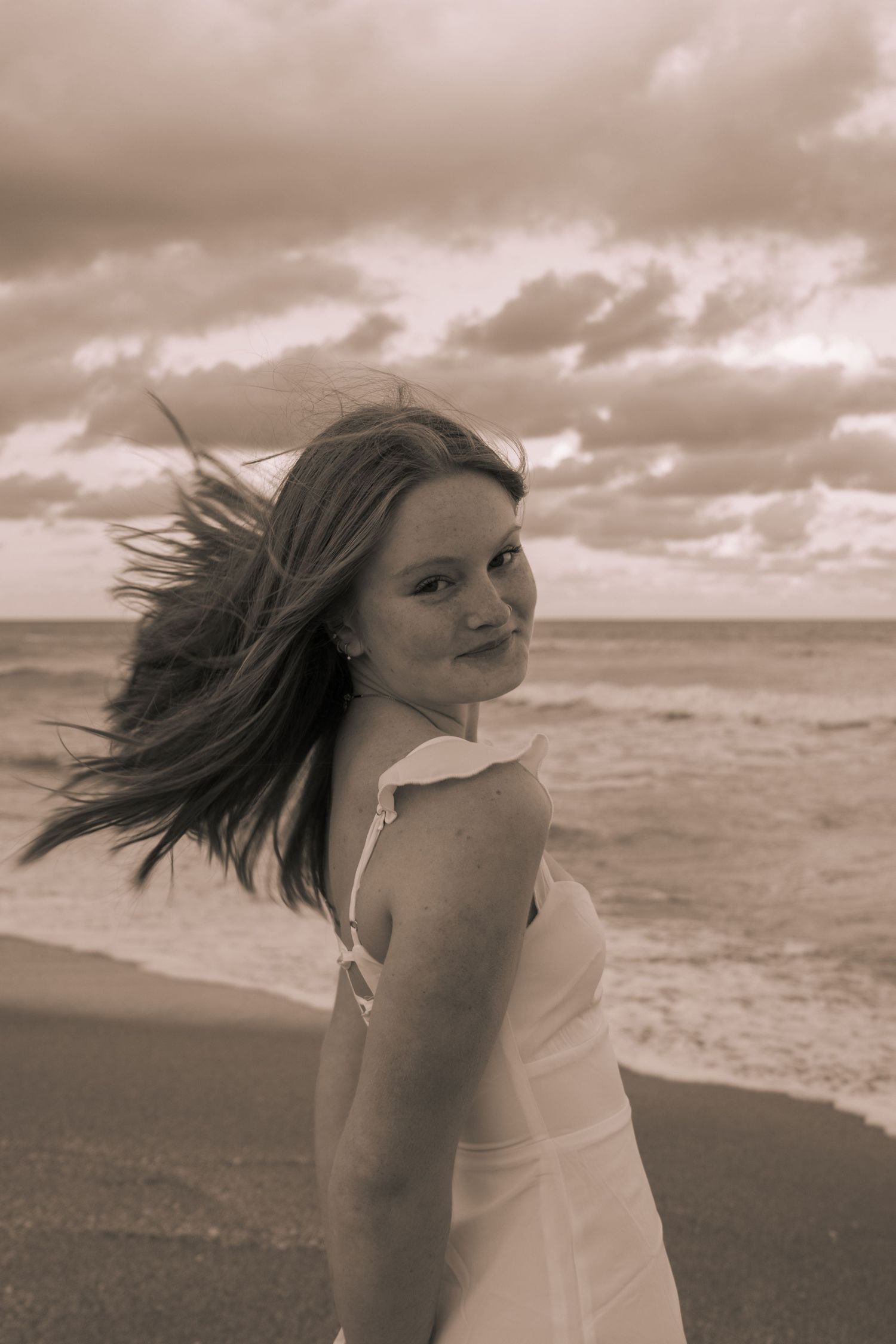 Sepia toned photo of young woman in white dress on beach with hair blowing in wind.