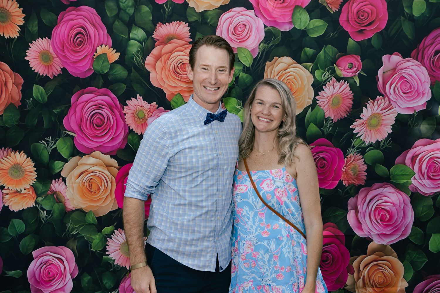 Man in bow tie and woman in floral dress smiling before a vibrant painted rose flower wall backdrop.
