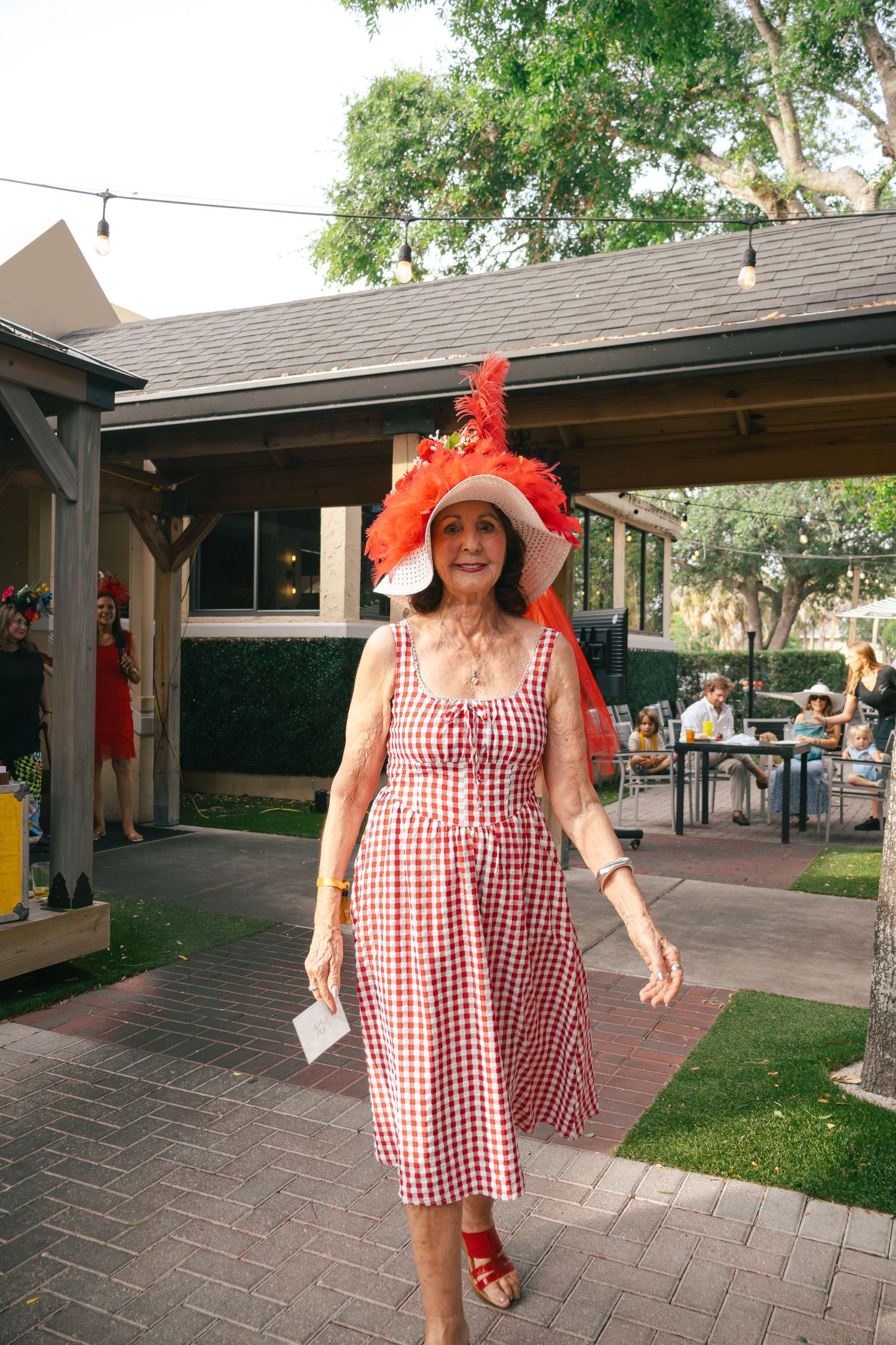 Woman in red gingham dress and large red feathered hat walks at Kentucky Derby party.