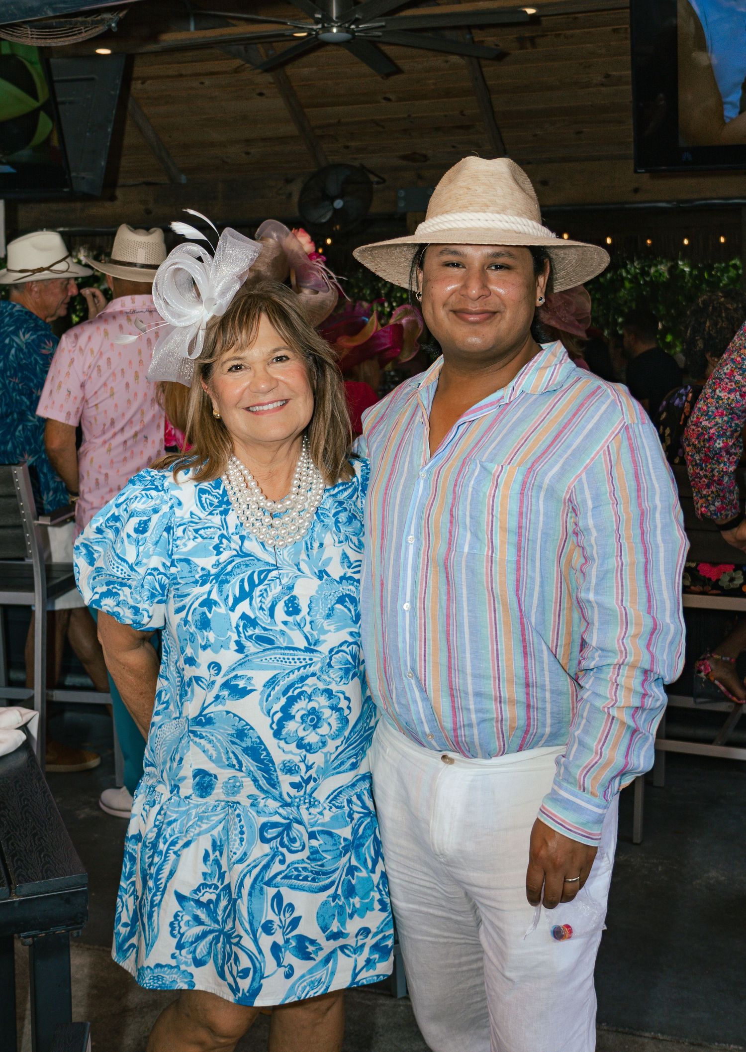 Woman in blue floral dress with white fascinator poses with man in striped shirt and panama hat.