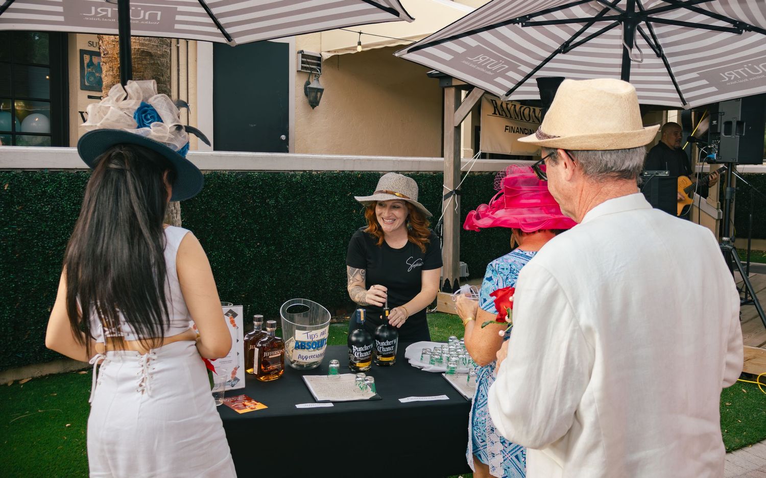 Guests in elegant hats browse a vendor table with bottles at a Kentucky Derby themed outdoor event.