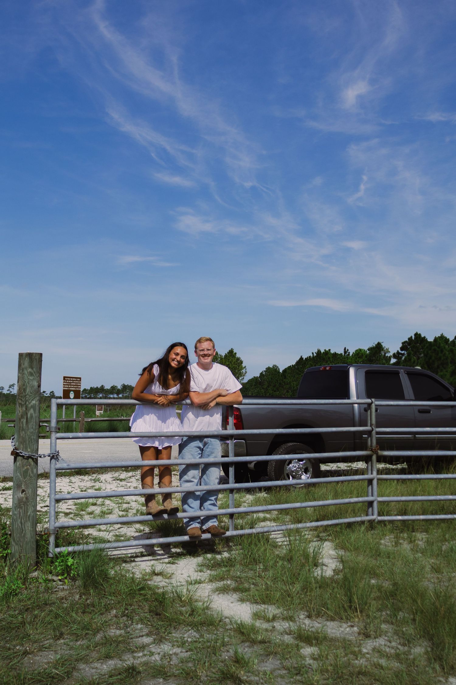 Smiling couple leaning on metal farm gate with pickup truck and blue sky in background.