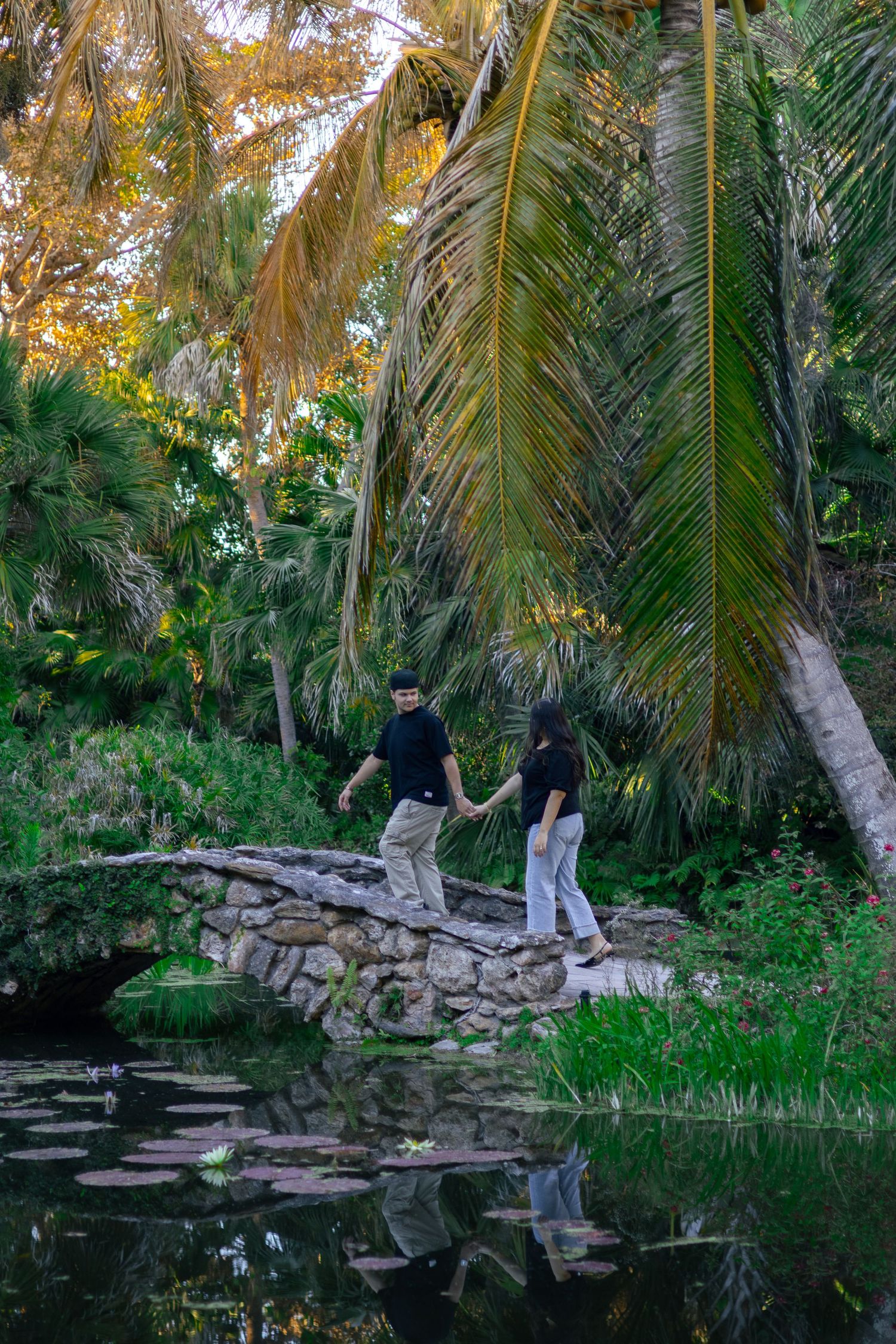 A couple walking hand in hand across a stone arch bridge reflected in a calm pond amid lush tropical greenery.