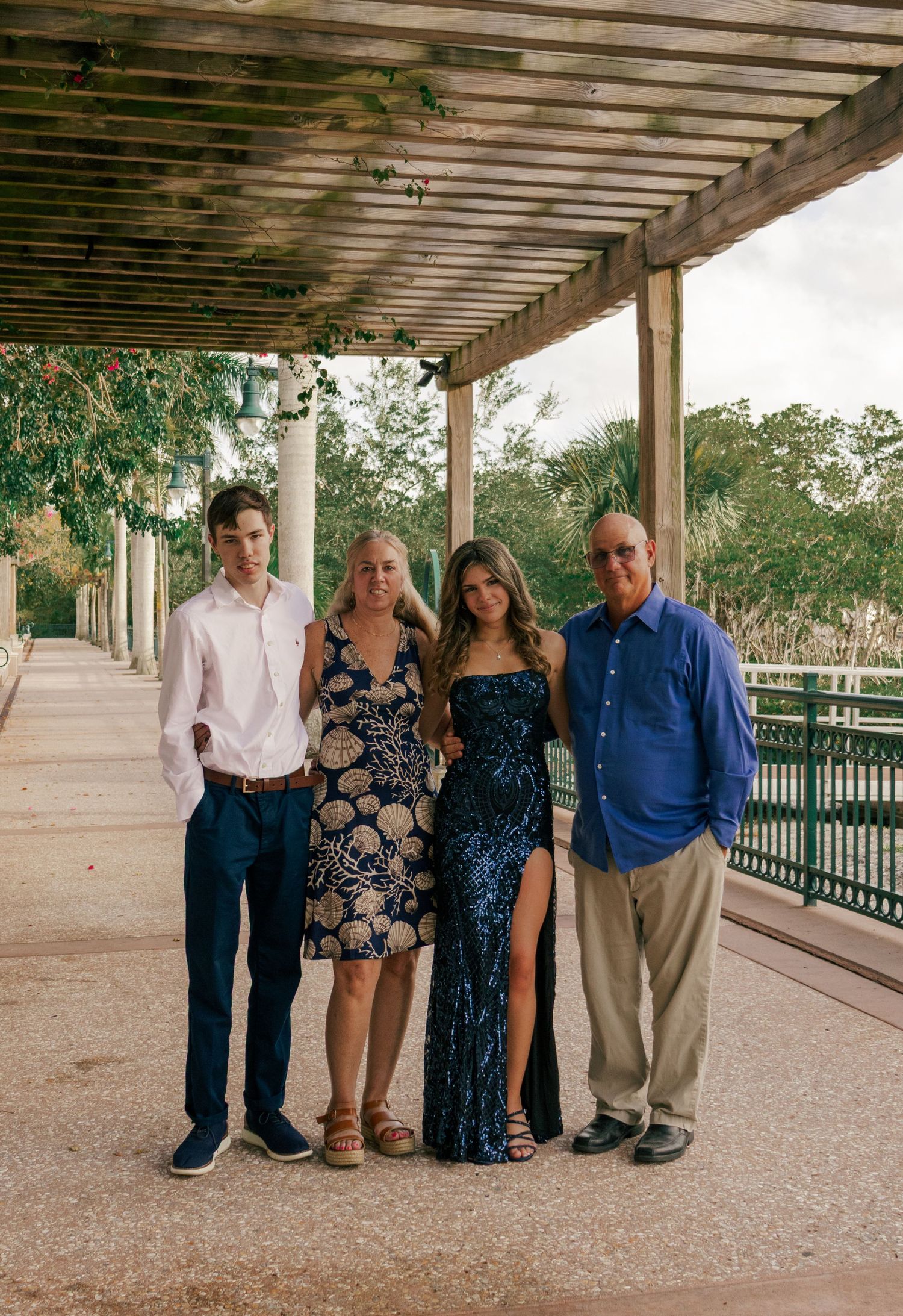 Family portrait of four people dressed formally under a wooden pergola in an outdoor park setting.