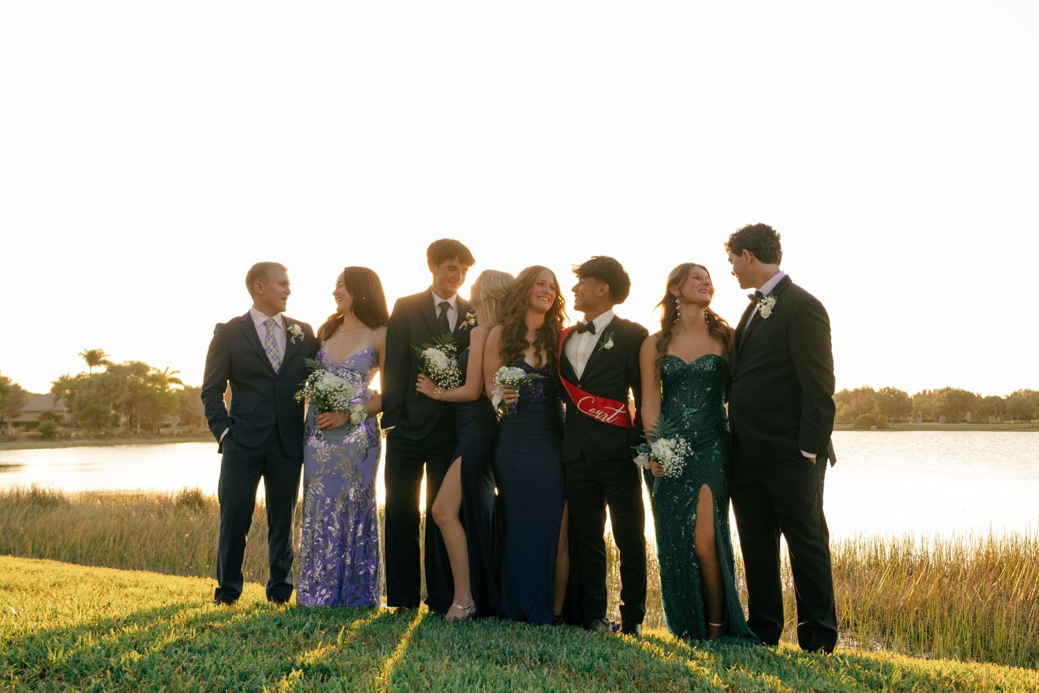 Seven smiling prom students in formal attire pose closely together by a serene lake at sunset.