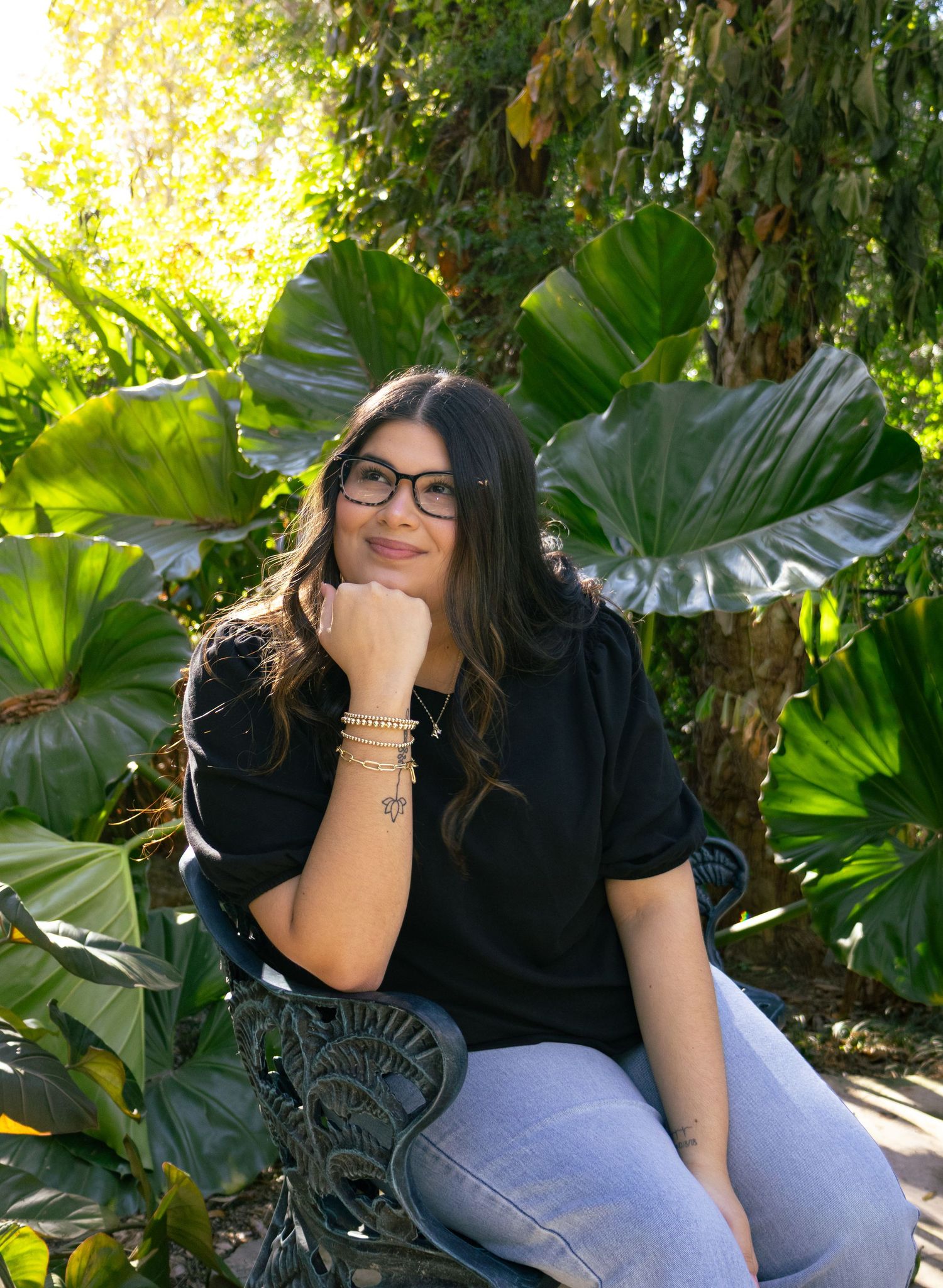 Smiling woman with glasses and bracelets poses on garden bench surrounded by large tropical leaves.