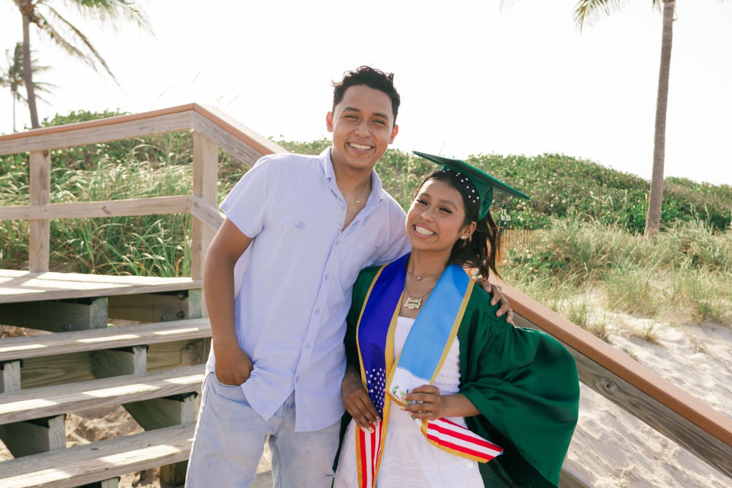 A smiling couple poses outdoors near a beach boardwalk, with the woman wearing a green graduation cap and gown.