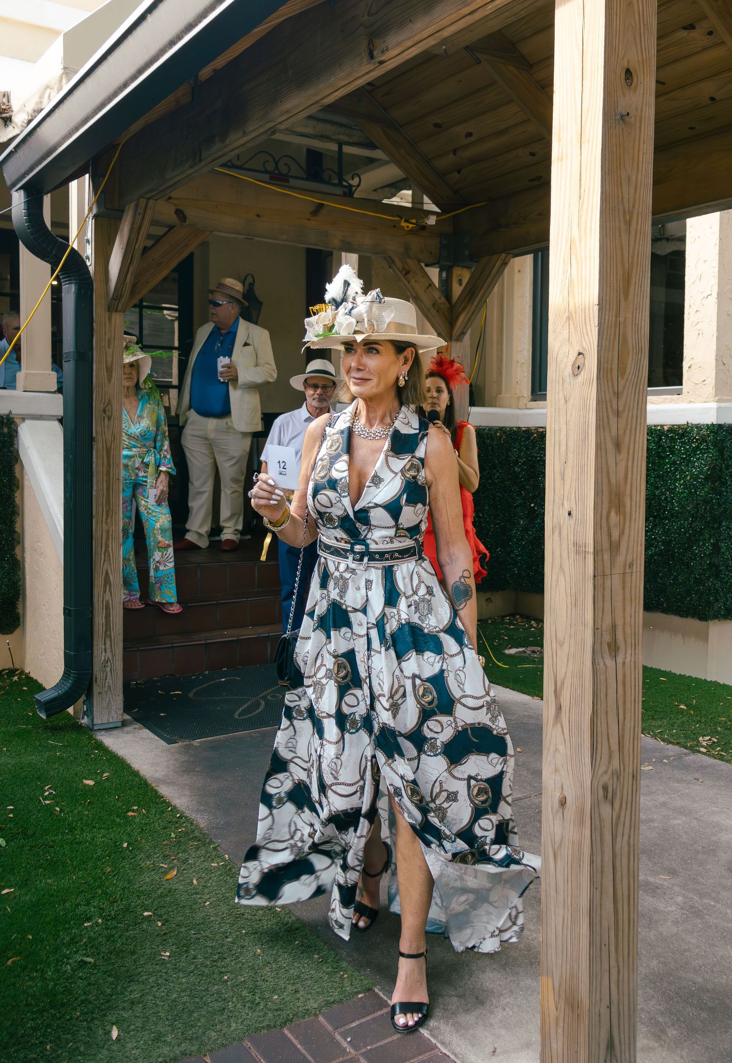 Woman in floral maxi dress and elaborate fascinator hat strolls through Derby party.
