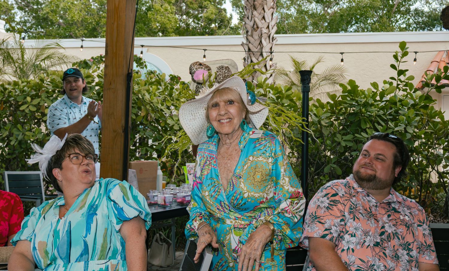 Three guests sit outdoors at a garden party, the center woman wearing an elegant white floral hat.