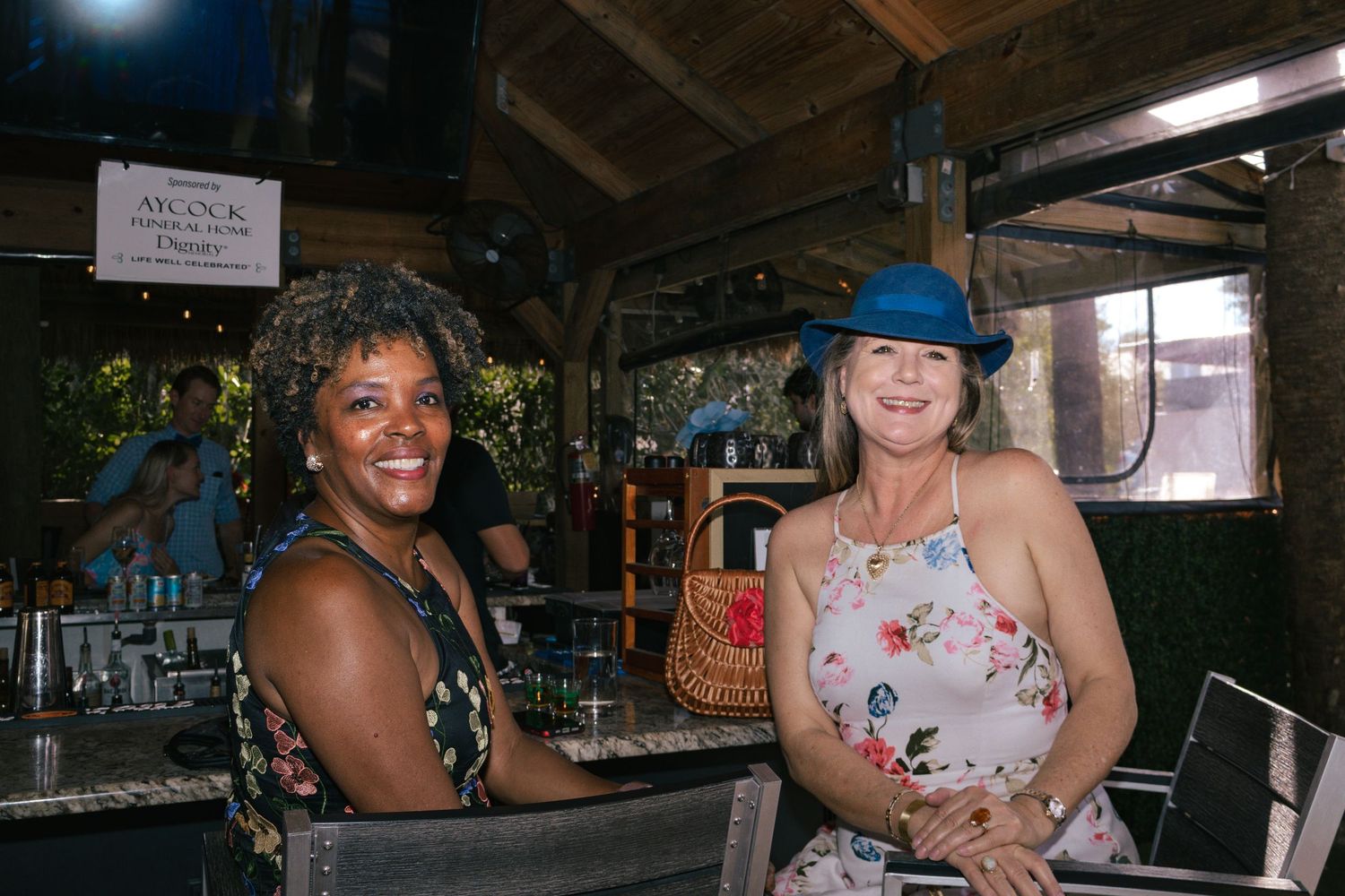 Two smiling women pose together at an outdoor bar, one wearing a blue hat and floral dress.