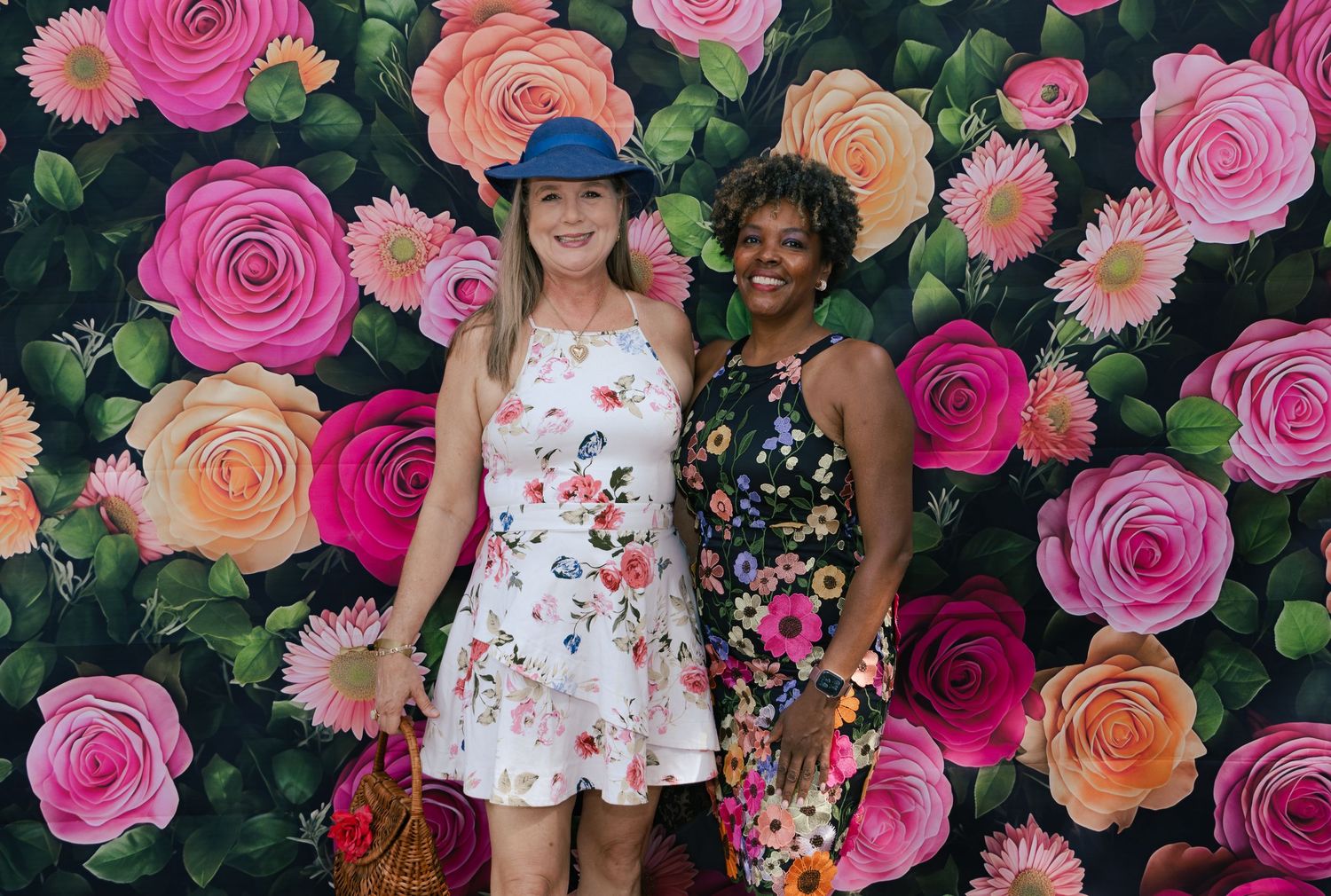 Two smiling women in summer dresses posing together in front of a vibrant painted pink and orange floral backdrop.