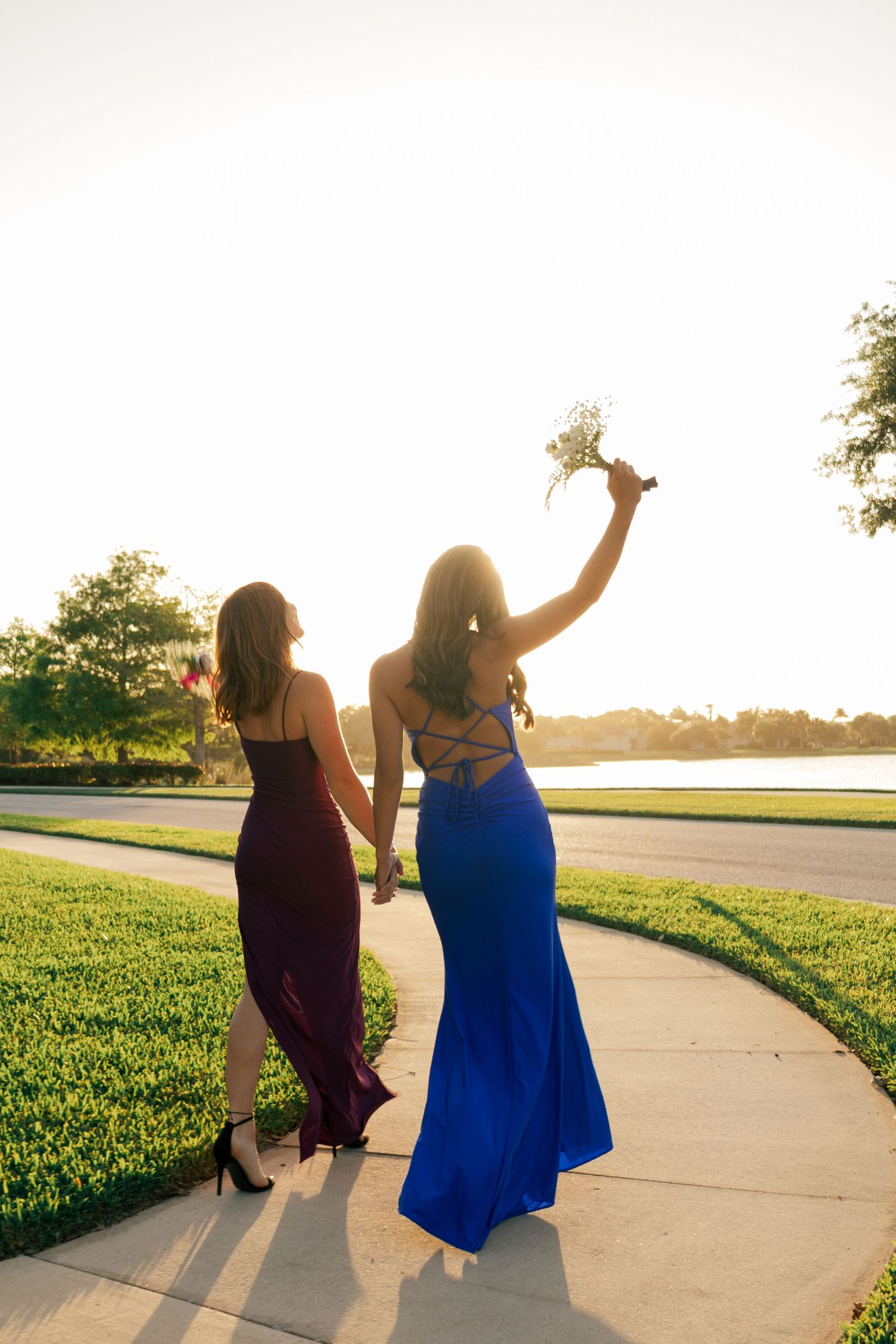 Two young women in blue and burgundy formal gowns walking at sunset, one raising a bouquet.