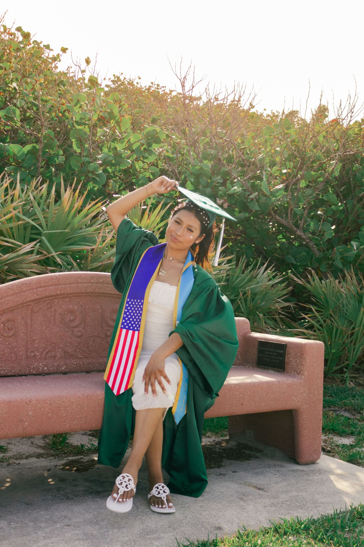 Female graduate in green cap and gown with American flag stole sits on pink stone bench outdoors.