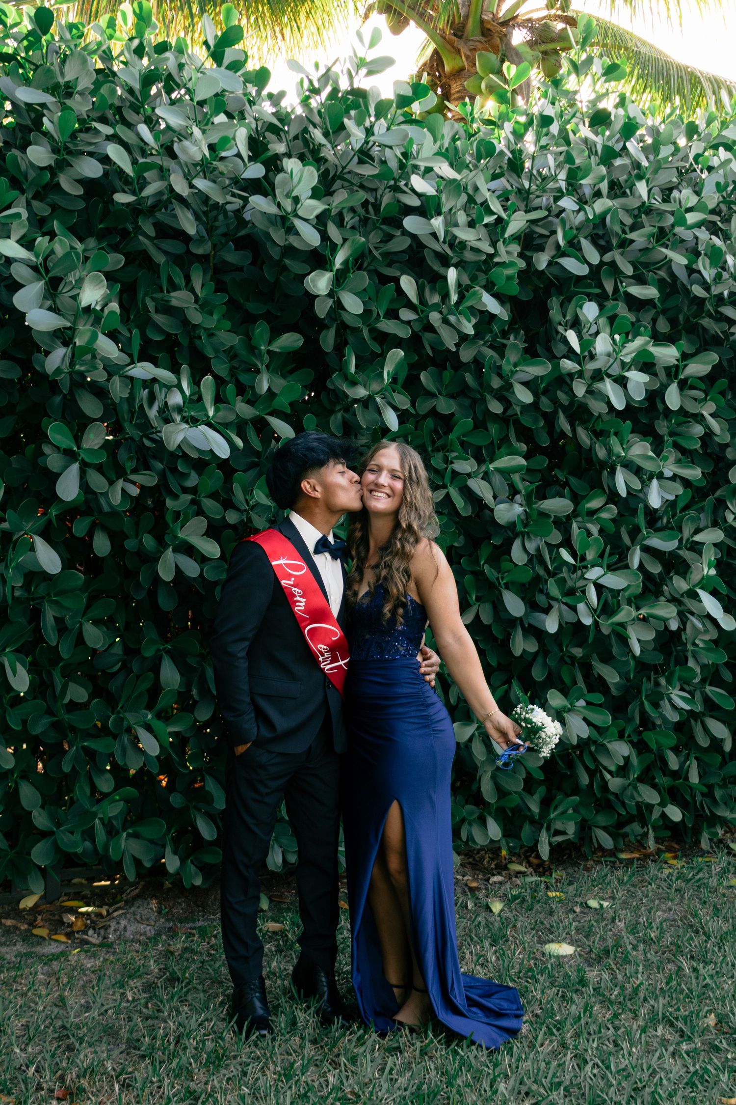 Prom couple poses against green hedge, young man in tuxedo with red sash kissing girls cheek.