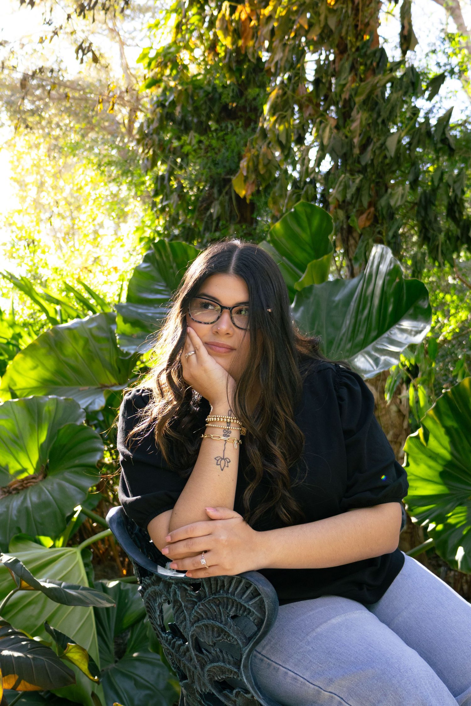 Young woman with glasses sitting among tropical plants, resting chin on hand in thoughtful pose outdoors.