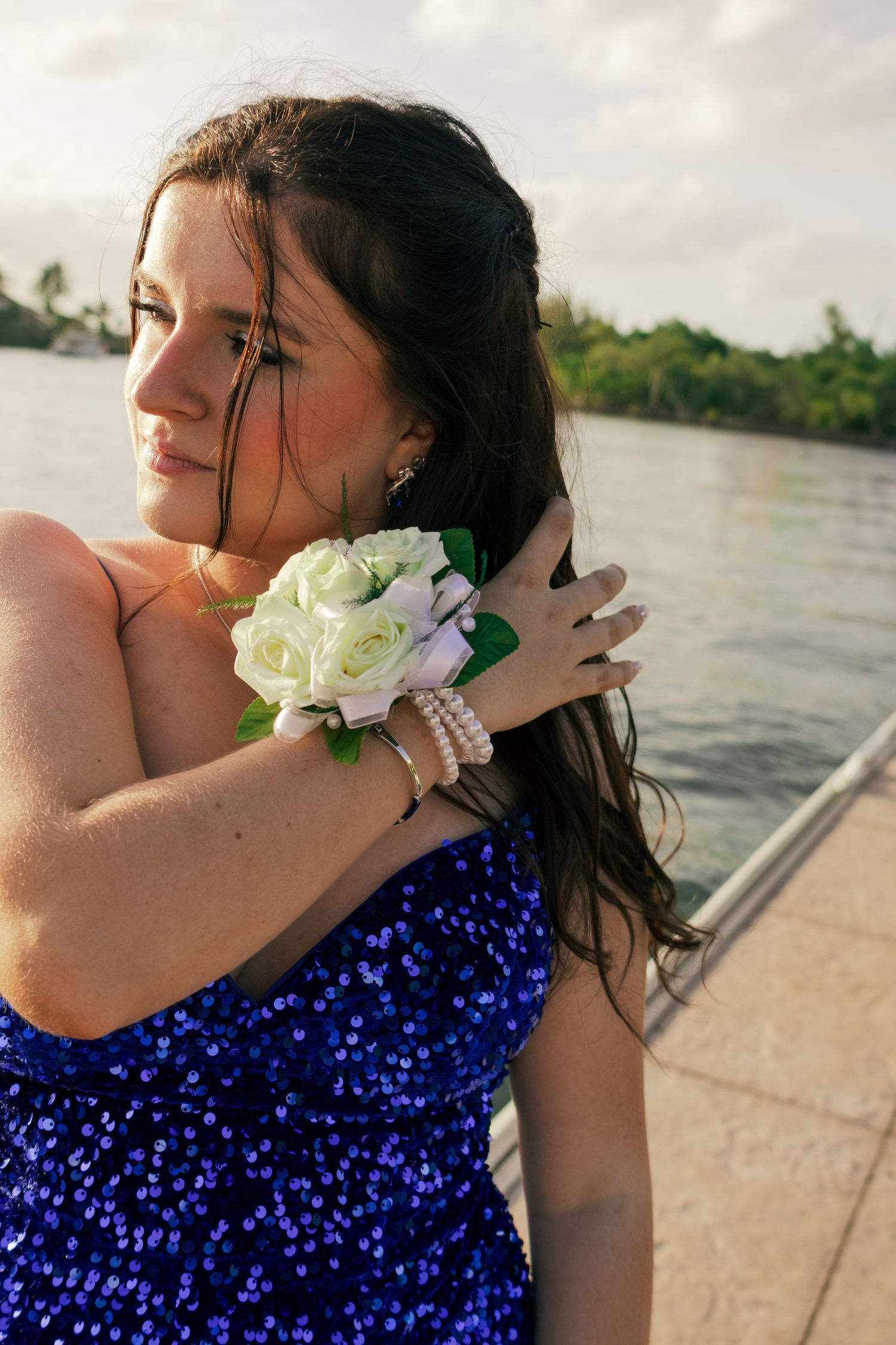Young woman in blue sequin dress with white rose wrist corsage poses by waterfront at golden hour.