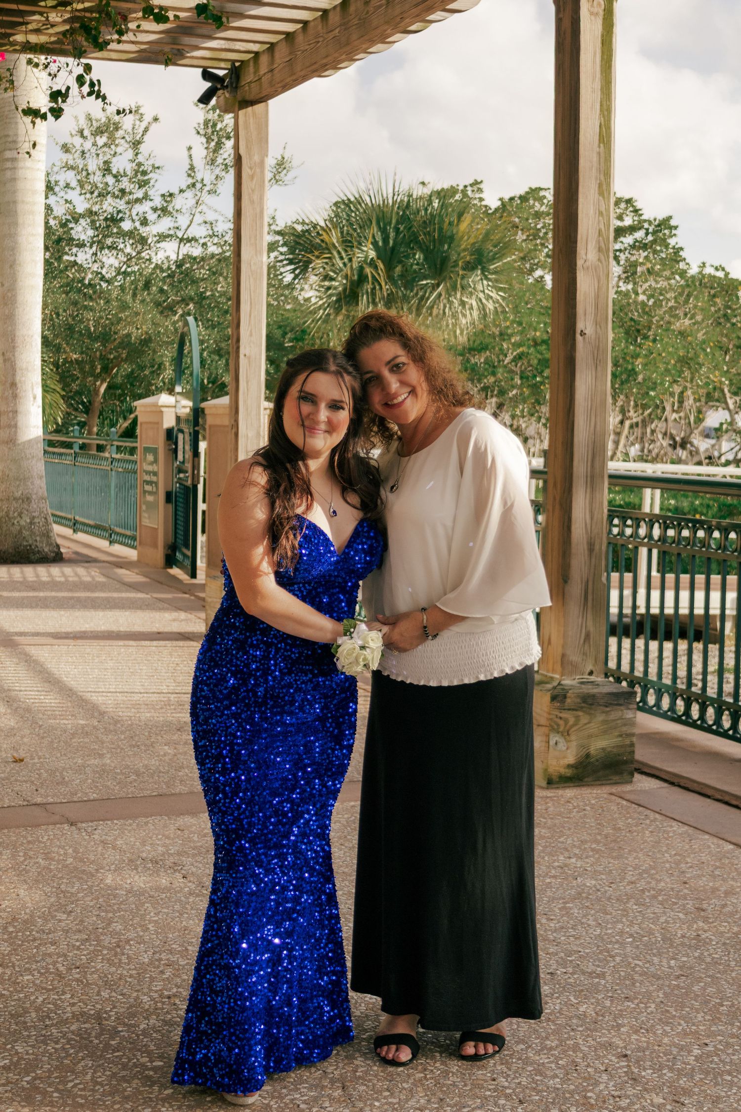 Young woman in blue sequin prom dress poses with older woman under outdoor pergola.