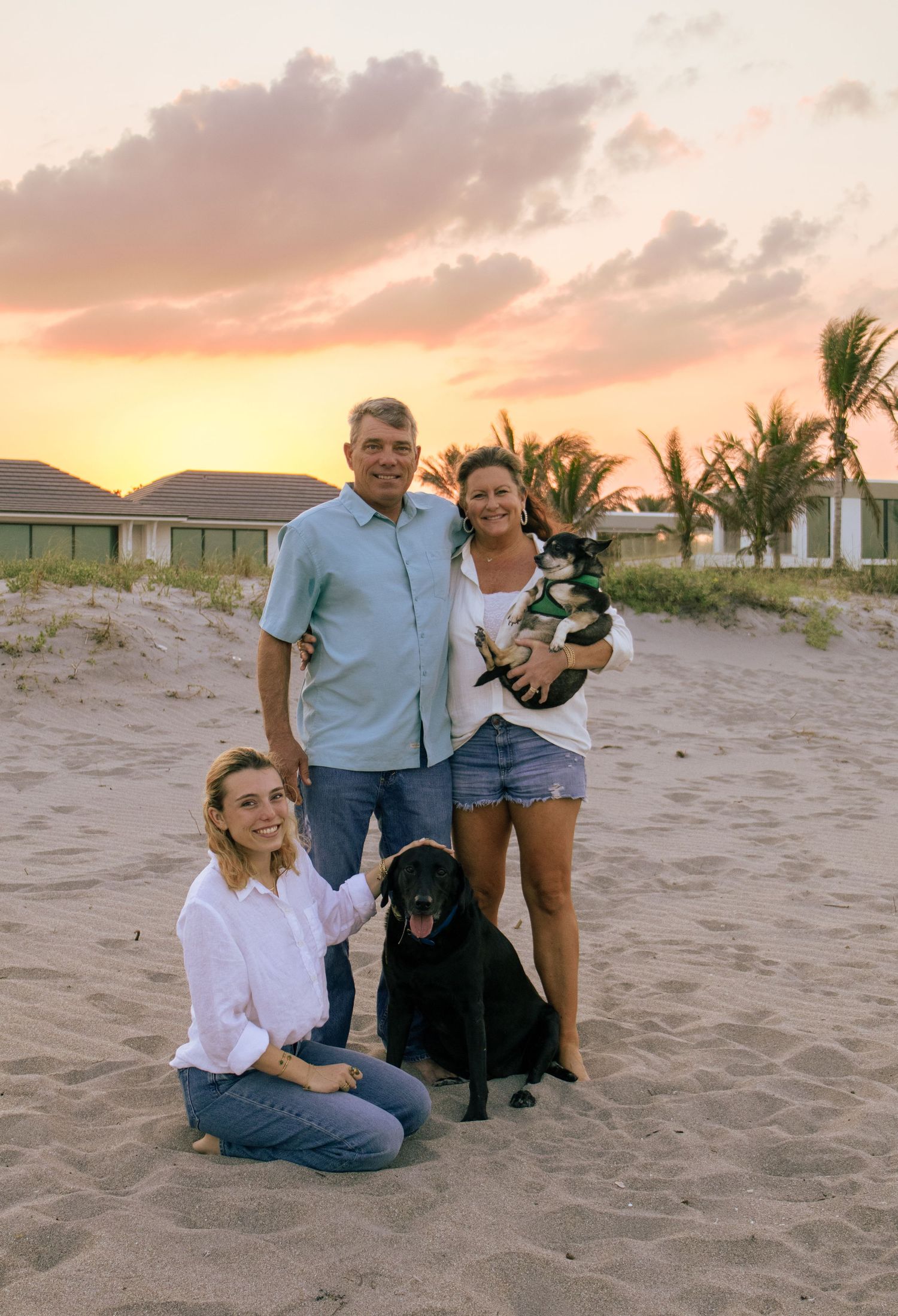 Family of three posing with two dogs on a sandy beach at golden sunset with palm trees in background.