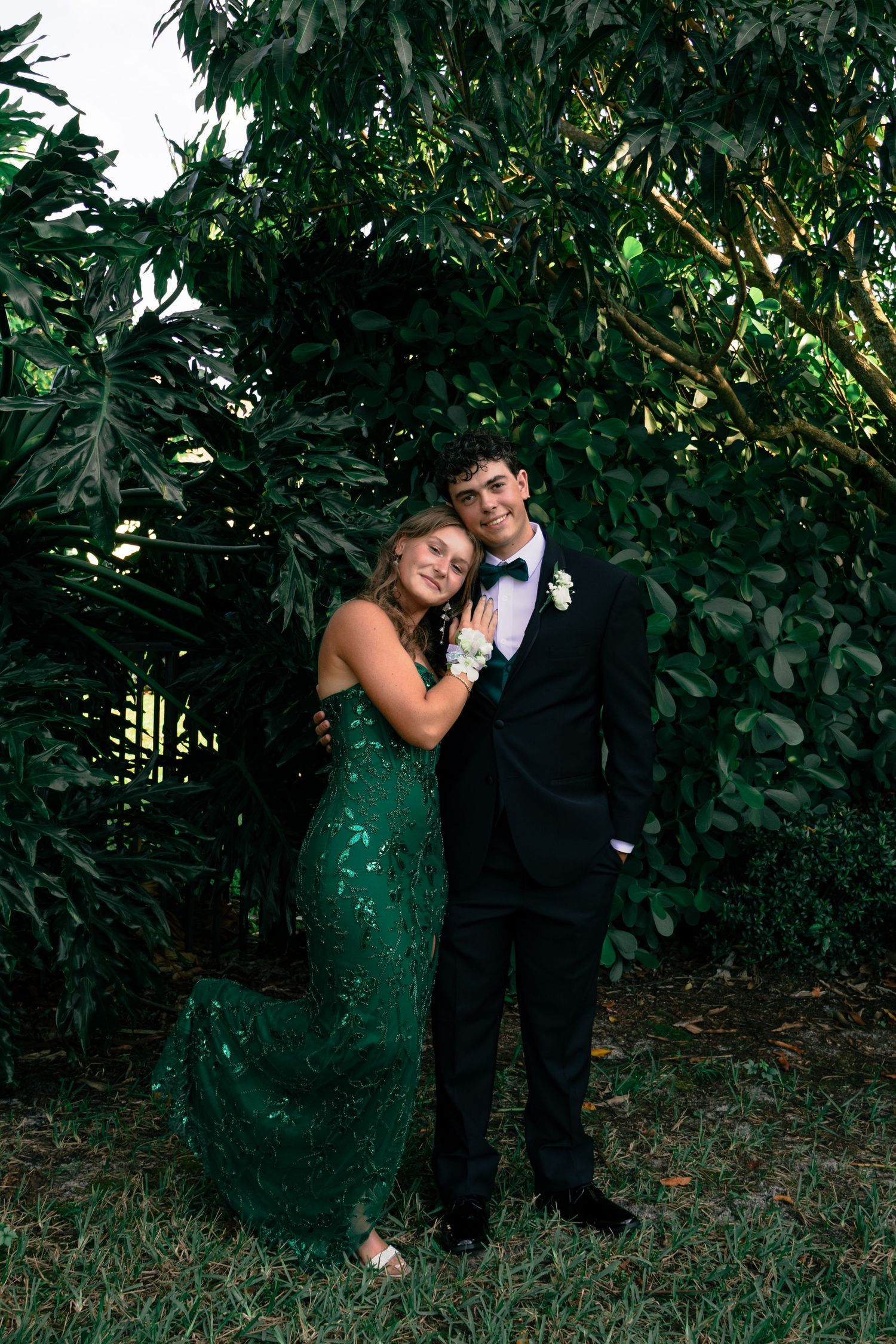 Prom couple embraces outdoors, girl in emerald green sequin gown and boy in classic black tuxedo.