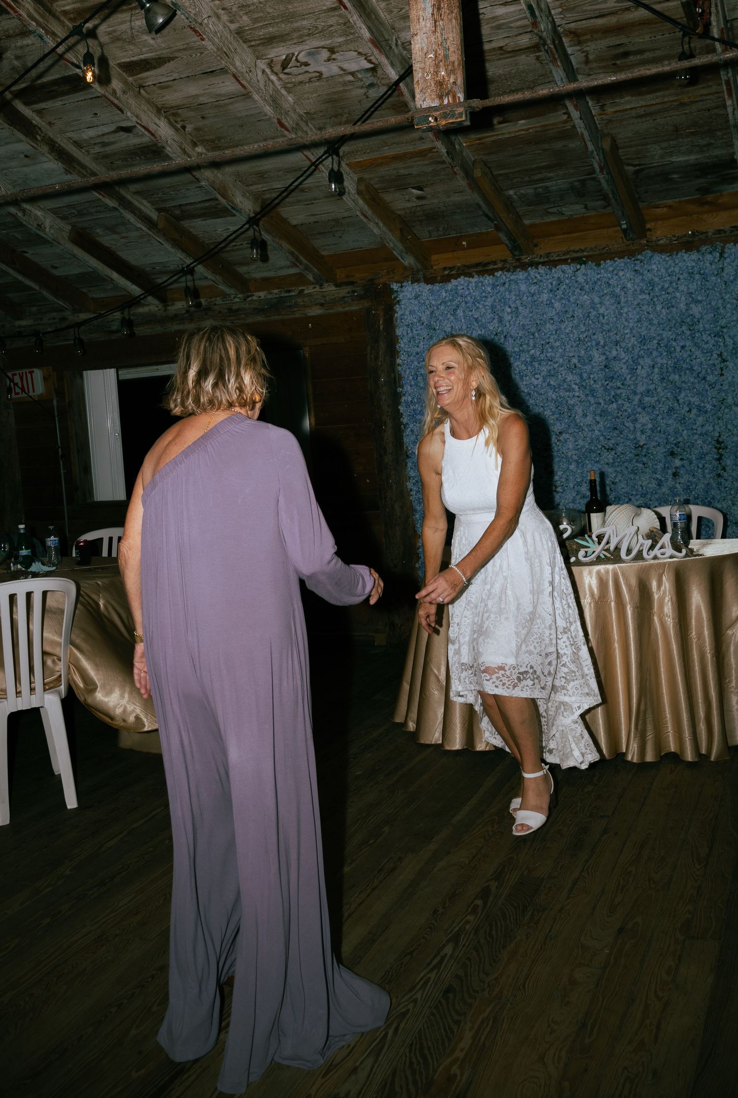 Bride in white lace dress dancing with a guest in lavender gown at rustic wedding venue.