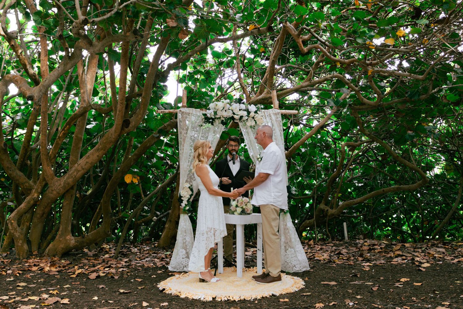 Bride and groom holding hands during an intimate outdoor wedding ceremony under a decorative arch surrounded by lush trees.