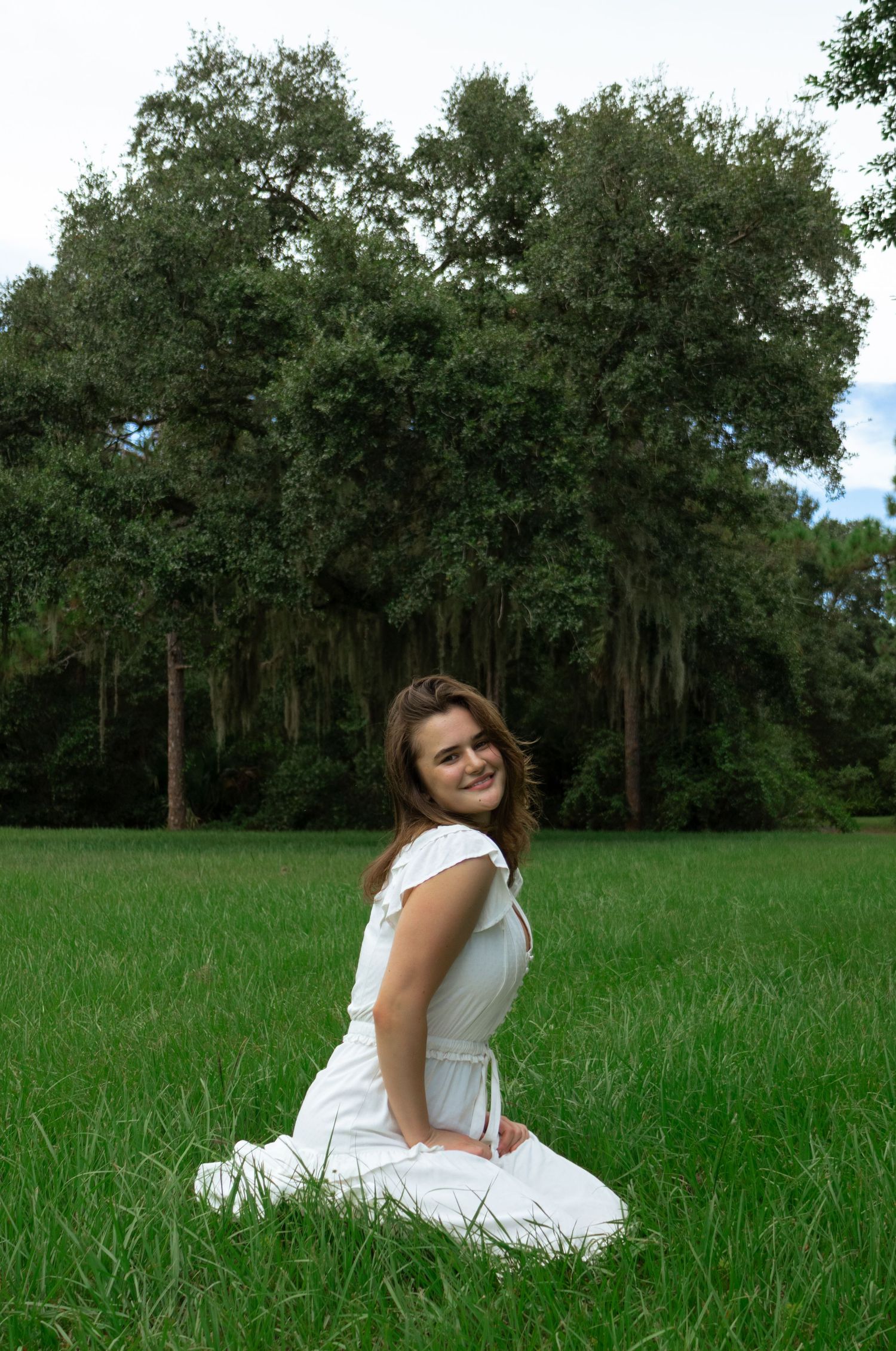 Young woman in white dress sitting on green grass field smiling, with large trees in background.