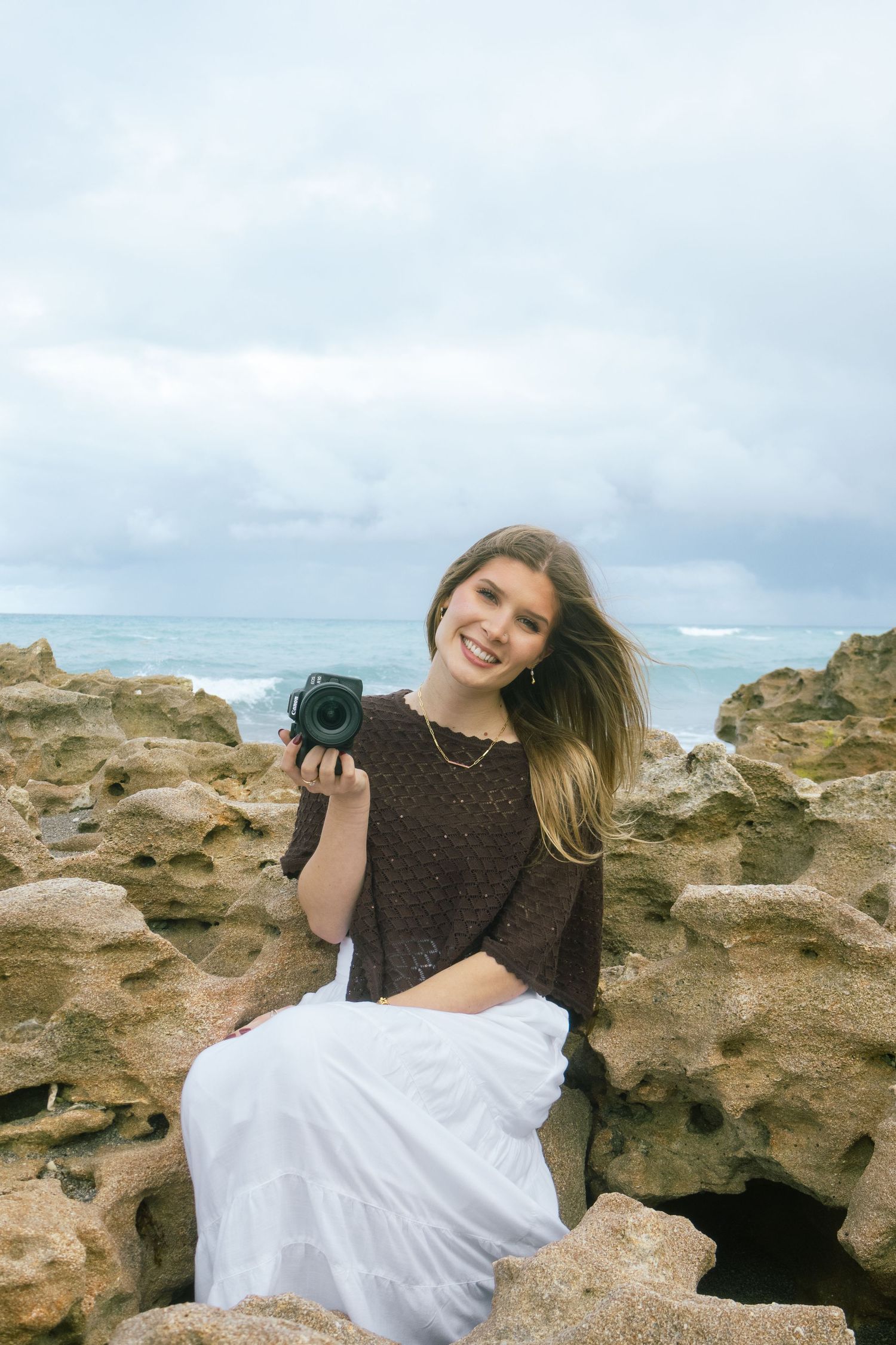 Smiling young female photographer sitting on rocks by the ocean holding a camera.