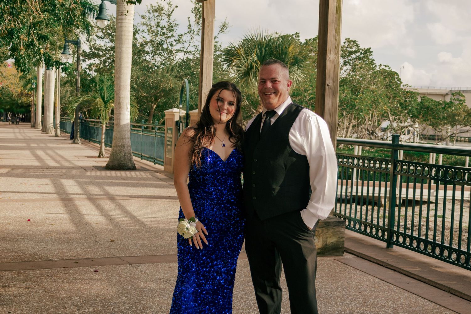 Couple posing in formal attire at outdoor venue, woman in blue sequin gown and man in black vest suit.