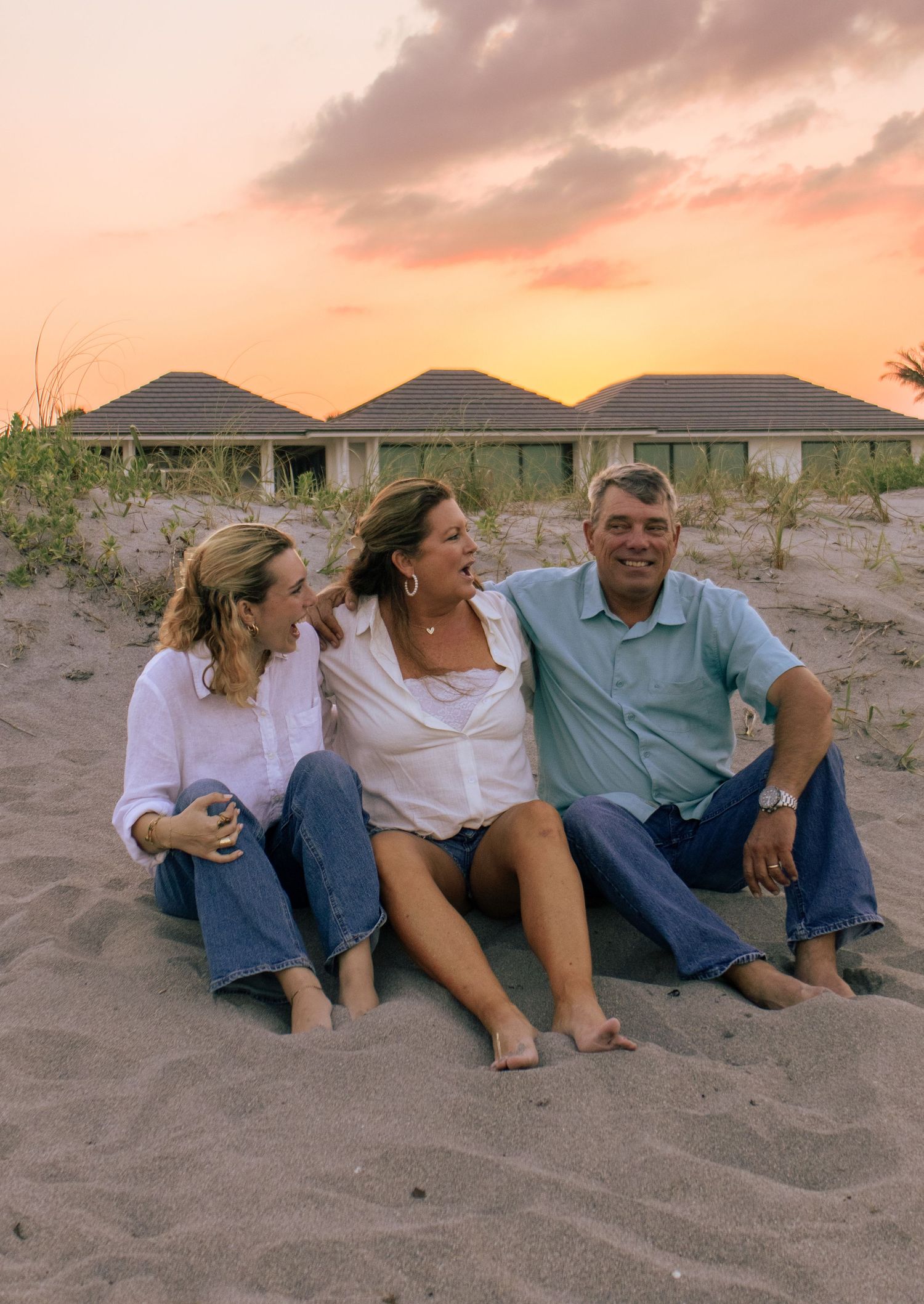 Family of three laughing together on sandy beach at sunset with beach houses in background.
