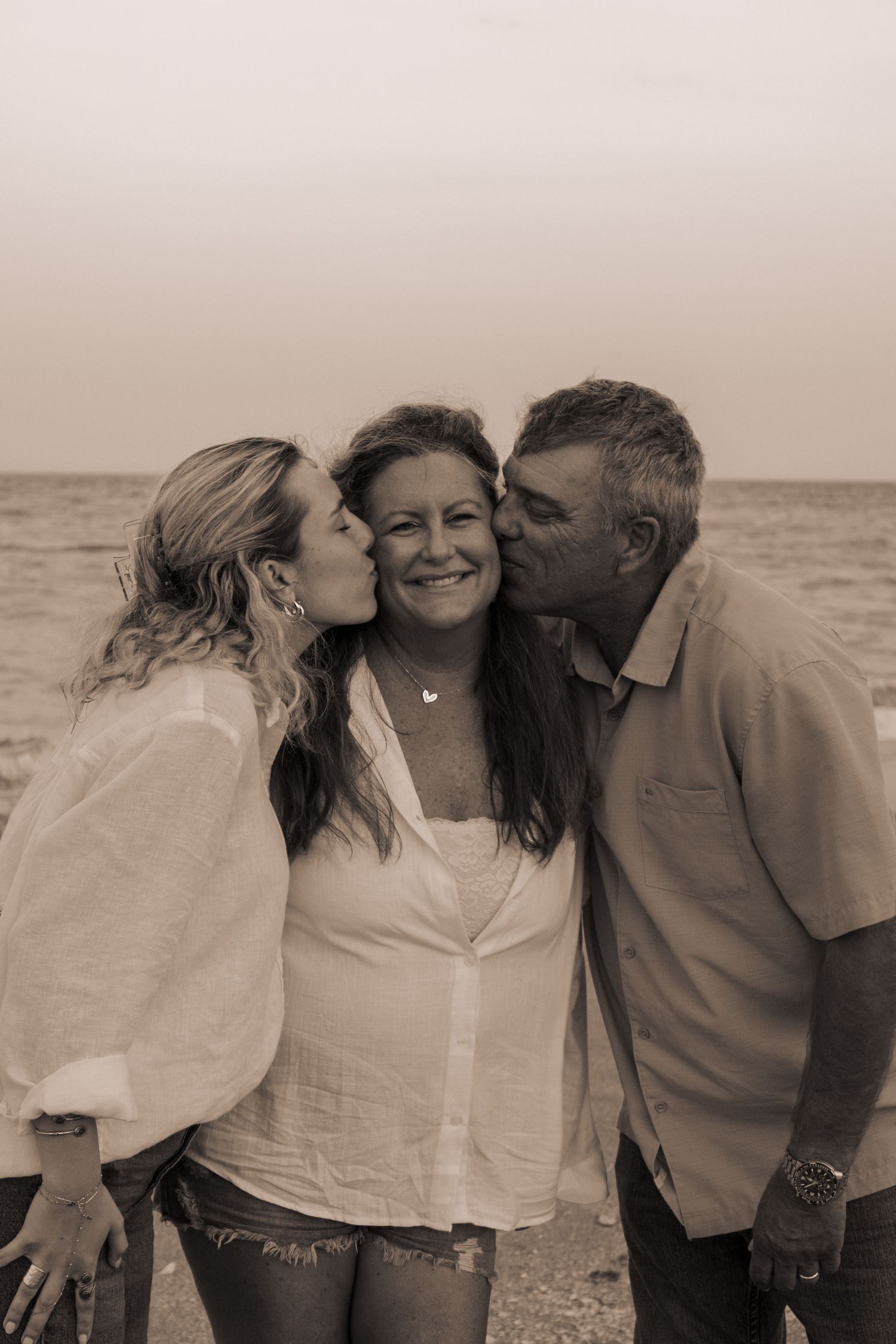 Sepia-toned photo of a smiling woman being kissed on cheeks by a younger woman and older man at a beach.