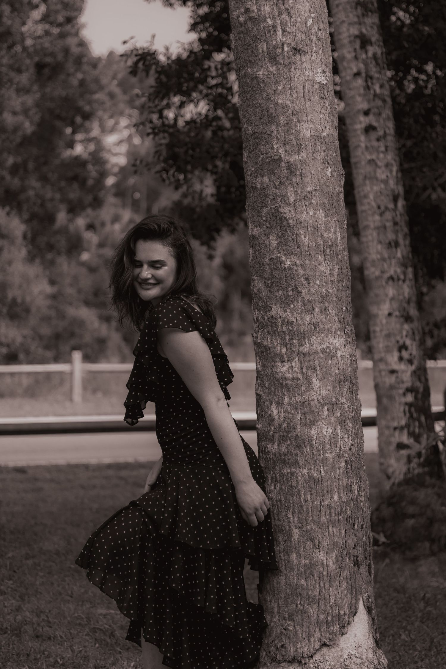 Young woman in black polka dot dress leaning against tree trunk in black and white outdoor photo.