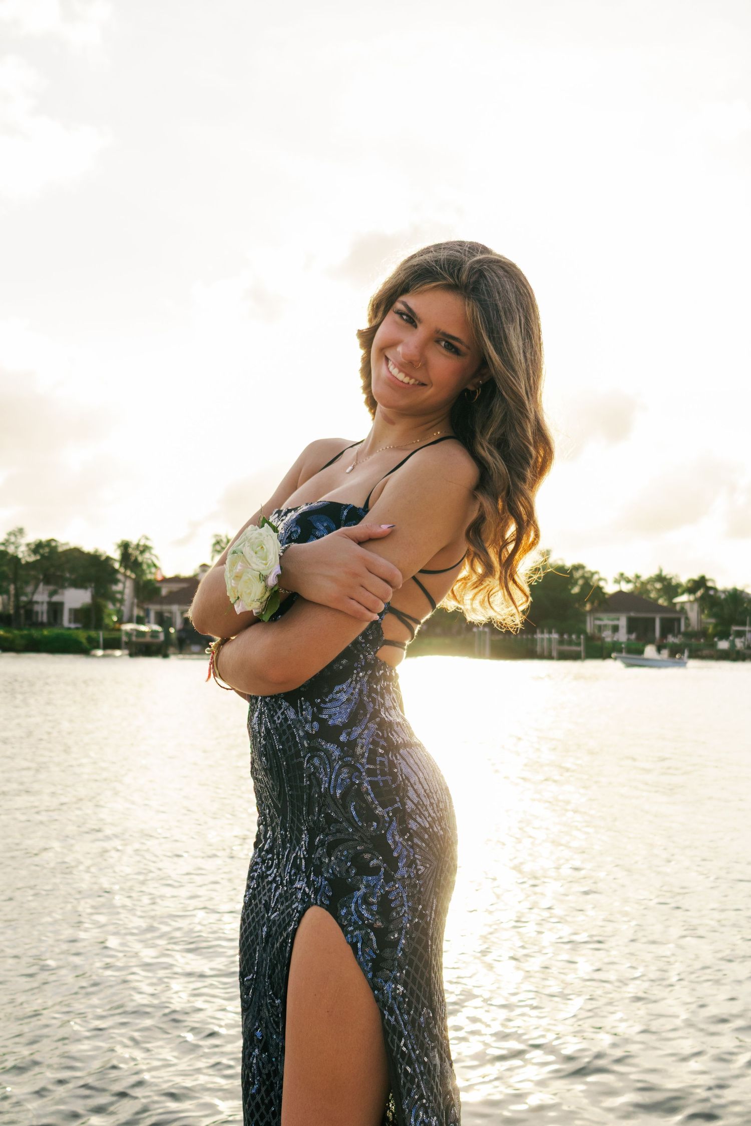 Young woman in blue sequin prom dress holding white corsage posing by the water at sunset.