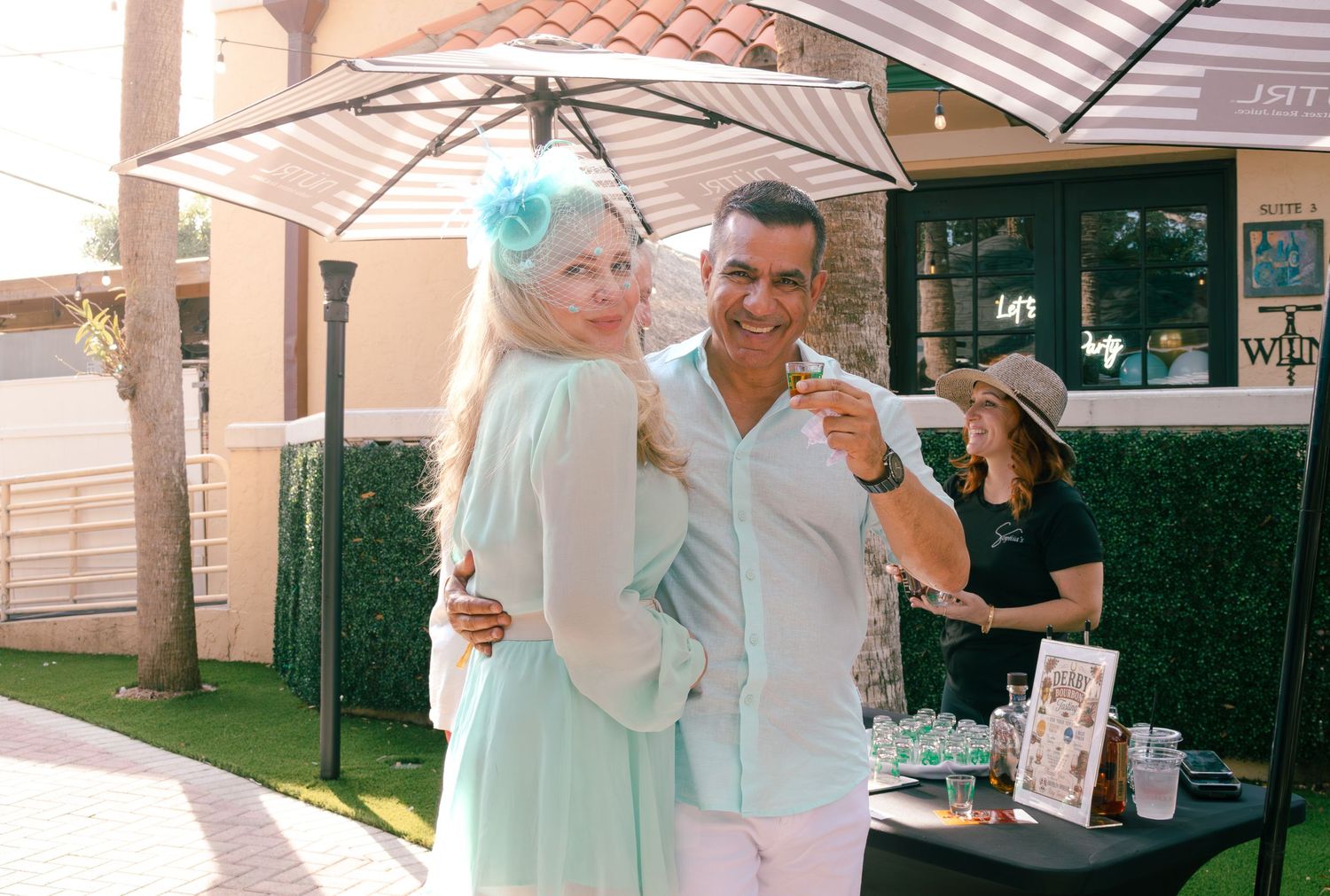 Couple poses together under striped umbrella at sunny outdoor social event.