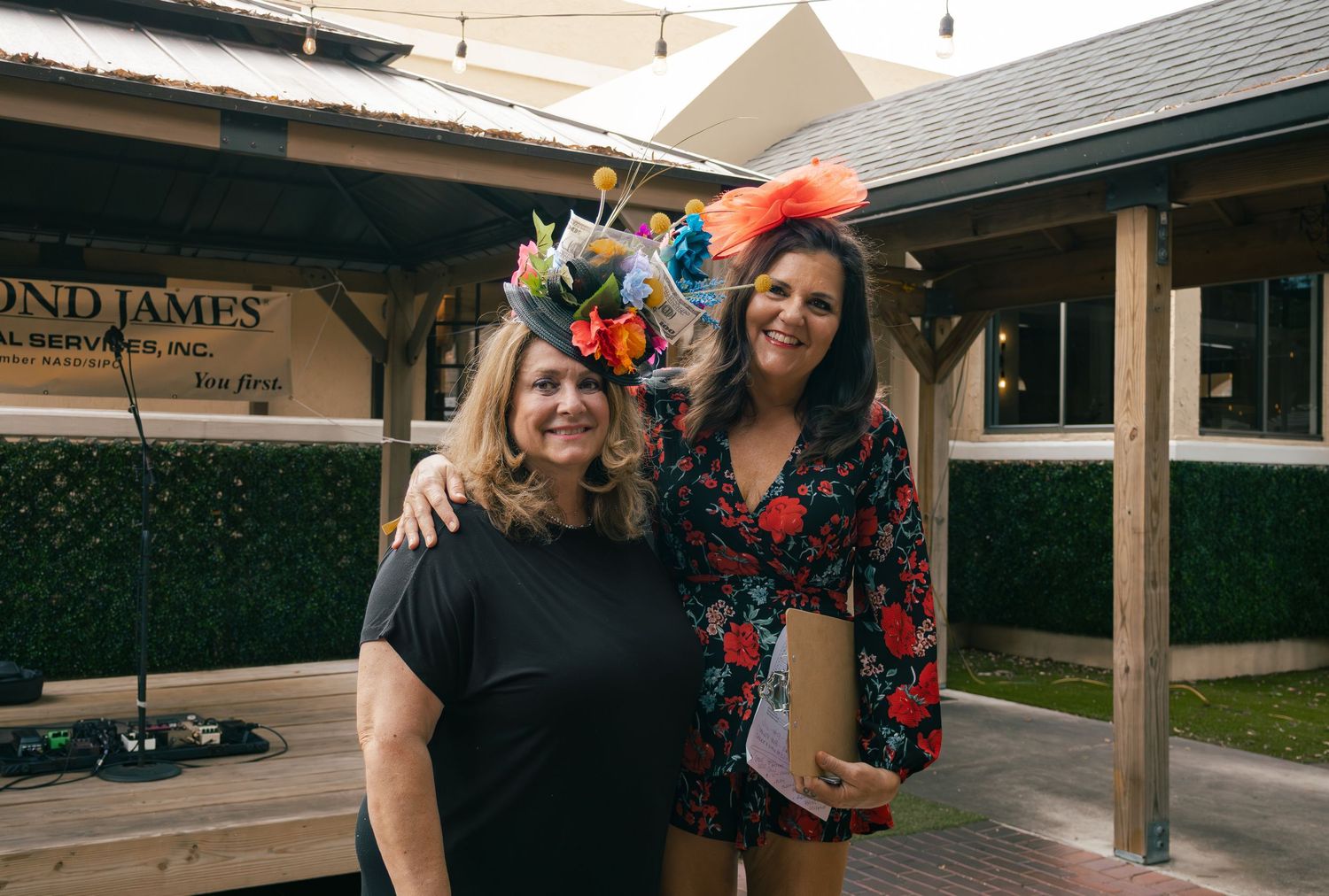 Two women wearing colorful floral fascinators smile together at an outdoor Kentucky Derby themed event.