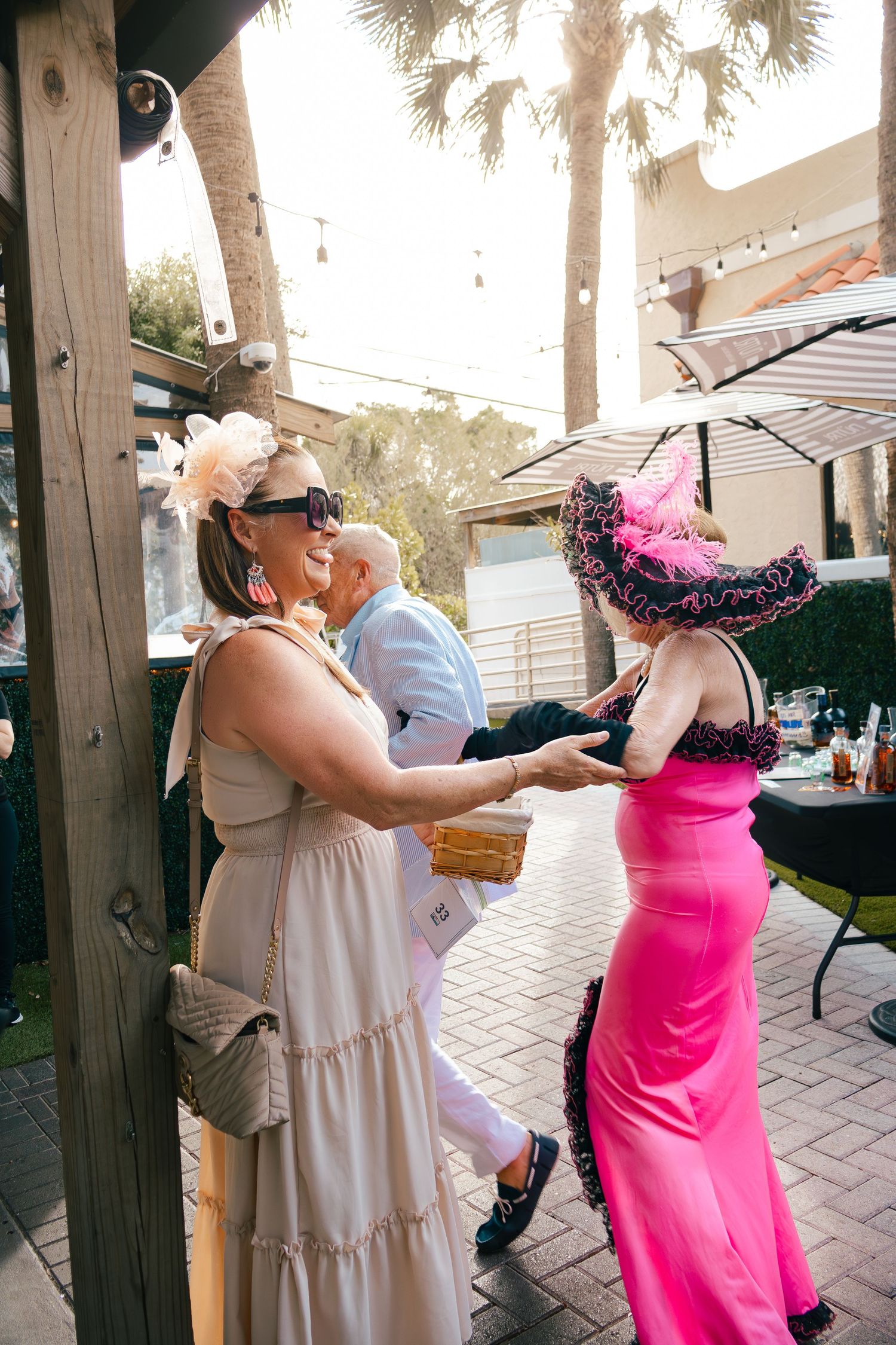 Women in elegant dresses and decorative hats socializing at outdoor Kentucky Derby themed party.