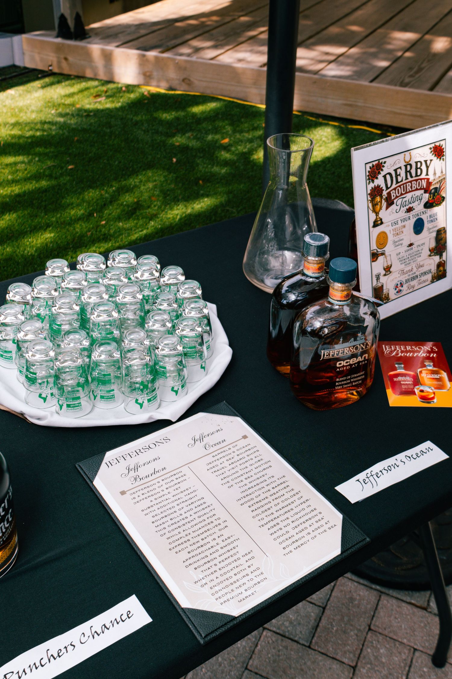 Jefferson's bourbon Derby cocktail station with recipe cards, shot glasses, and whiskey bottles on a black draped table outdoors.