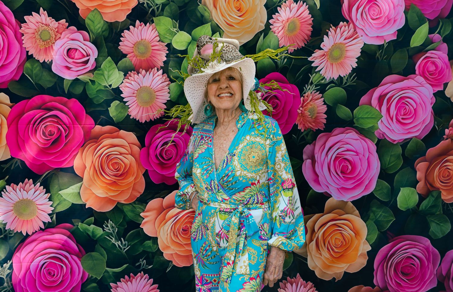 Elegant senior woman in turquoise floral dress and white hat posing before a vibrant painted rose flower wall backdrop.