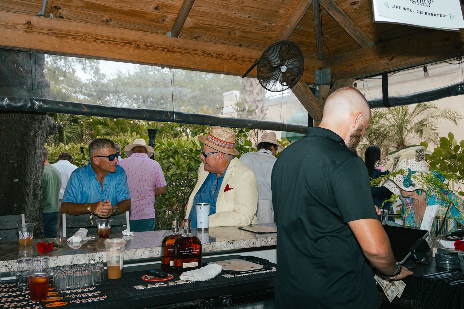 Guests mingle at an outdoor bar with bourbon bottles, enjoying a casual social gathering.