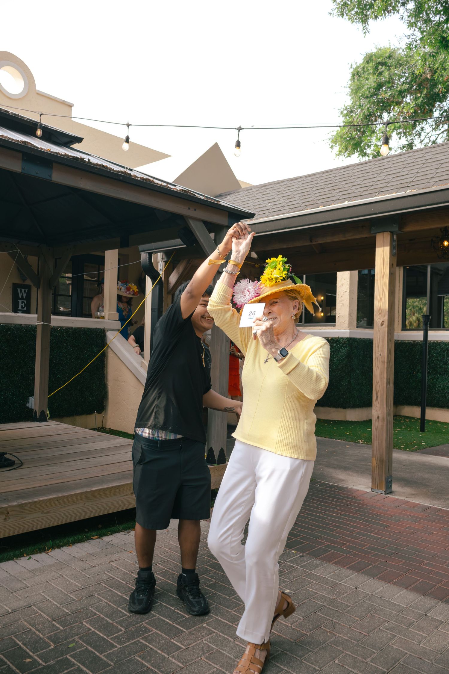 Elderly woman in yellow sweater and floral hat dancing with man in black outfit at outdoor venue.