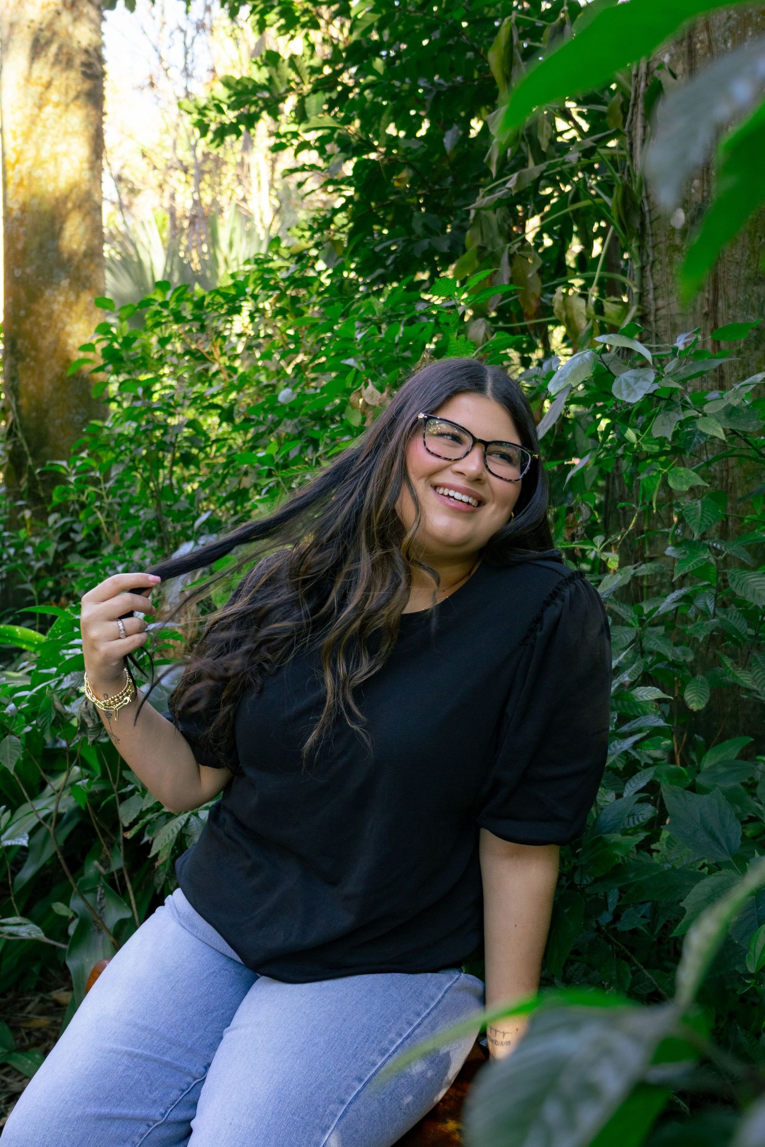 Smiling woman with glasses in black top plays with hair while sitting in green garden setting.